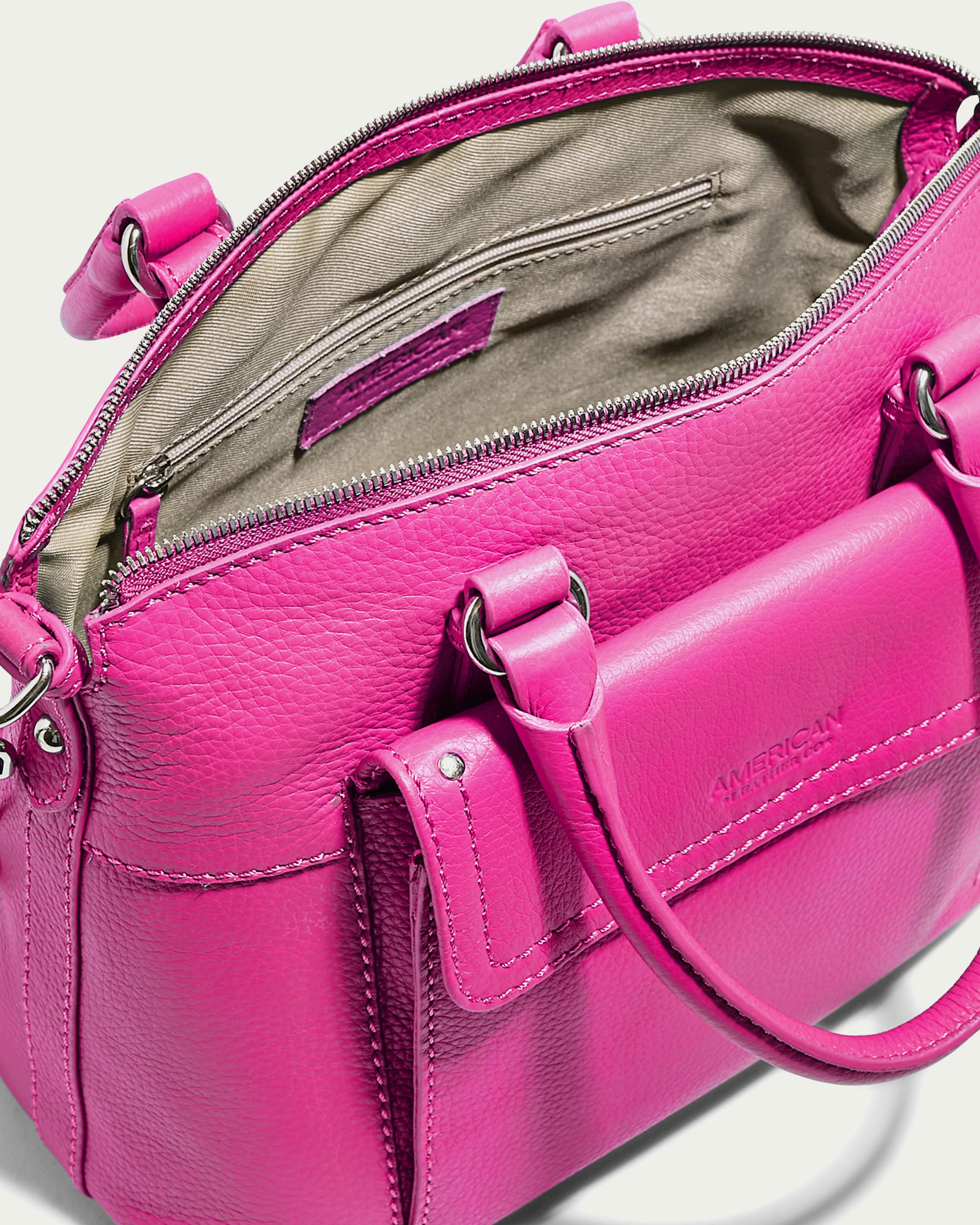 A vibrant pink genuine leather handbag with an open top, revealing a beige fabric interior. The Vona Convertible Dome Satchel from American Leather Co. showcases metal hardware, a zippered pocket inside, and a front external pocket. The handles are attached with metal rings, and the brand name "American Leather Co." is embossed on the front pocket.