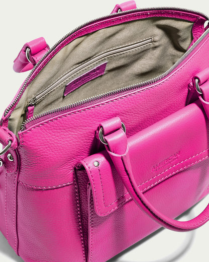 A vibrant pink genuine leather handbag with an open top, revealing a beige fabric interior. The Vona Convertible Dome Satchel from American Leather Co. showcases metal hardware, a zippered pocket inside, and a front external pocket. The handles are attached with metal rings, and the brand name "American Leather Co." is embossed on the front pocket.