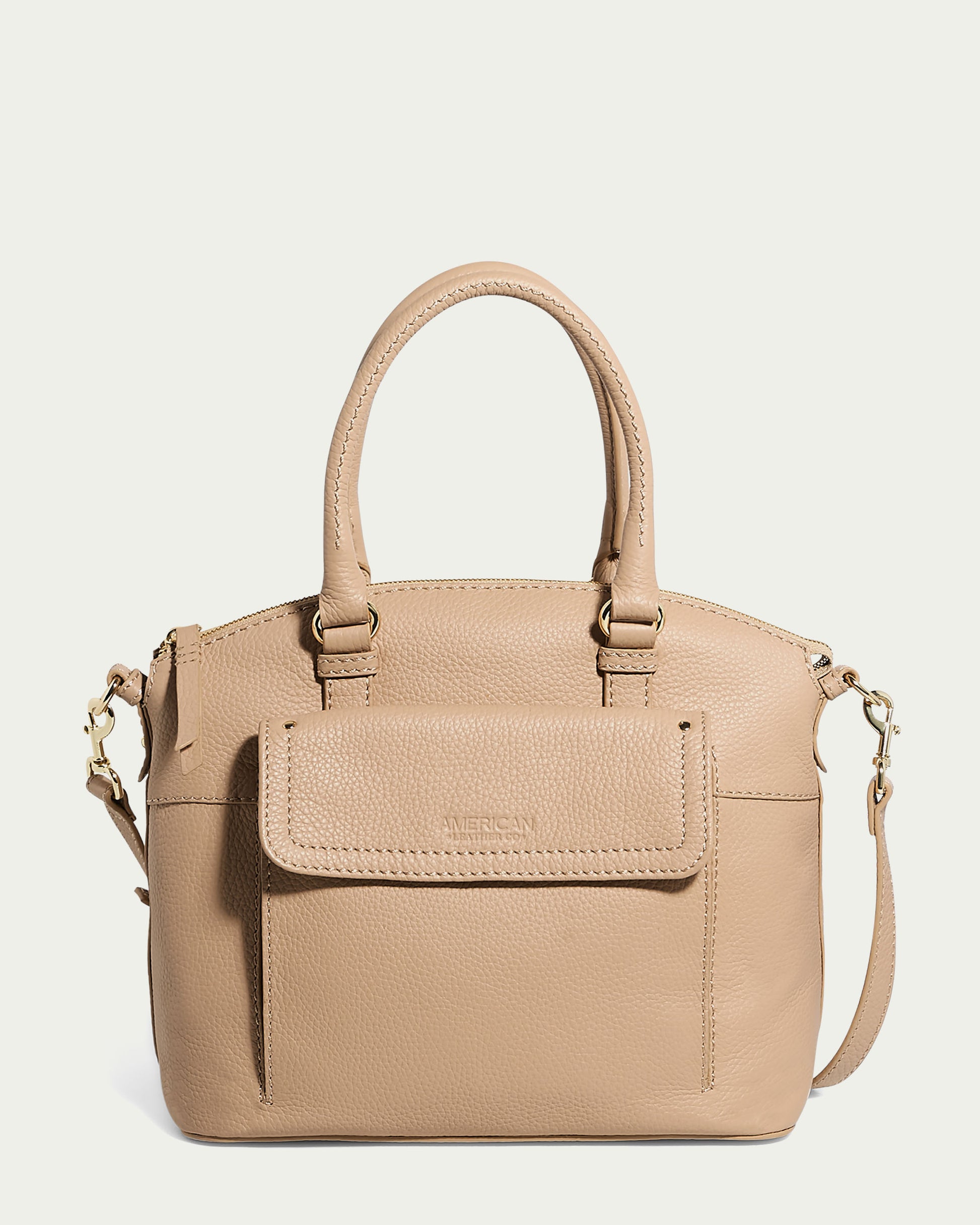 A macadamia Forever Pebble leather Vona Convertible Dome Satchel by American Leather Co. with dual top handles and a detachable shoulder strap. The handbag features a front flap pocket embossed with text and a zippered main compartment. The material has a pebbled texture, and the hardware appears to be gold-toned.