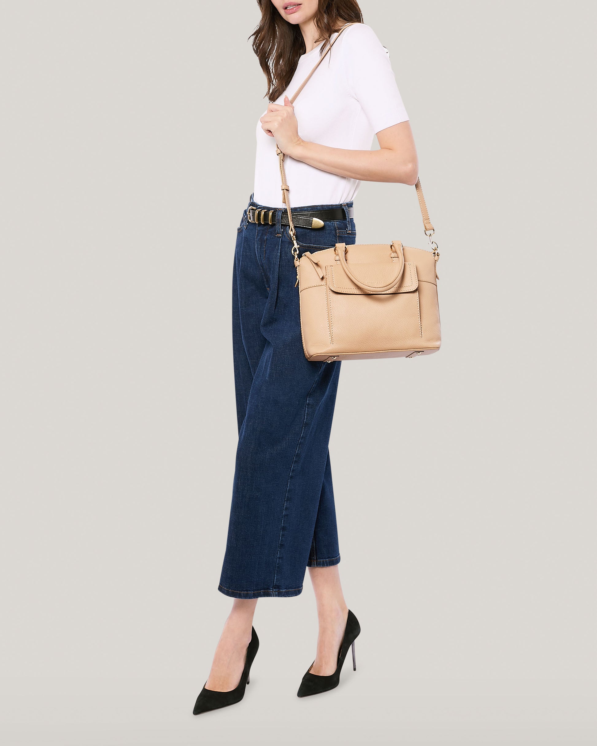 A woman is posing against a light-colored background. She is wearing a white T-shirt, high-waisted blue wide-legged jeans, and black heels. She carries the beige Vona Convertible Dome Satchel from American Leather Co., which features a shoulder strap. Her head is turned slightly, and her hair falls loosely over her shoulder.