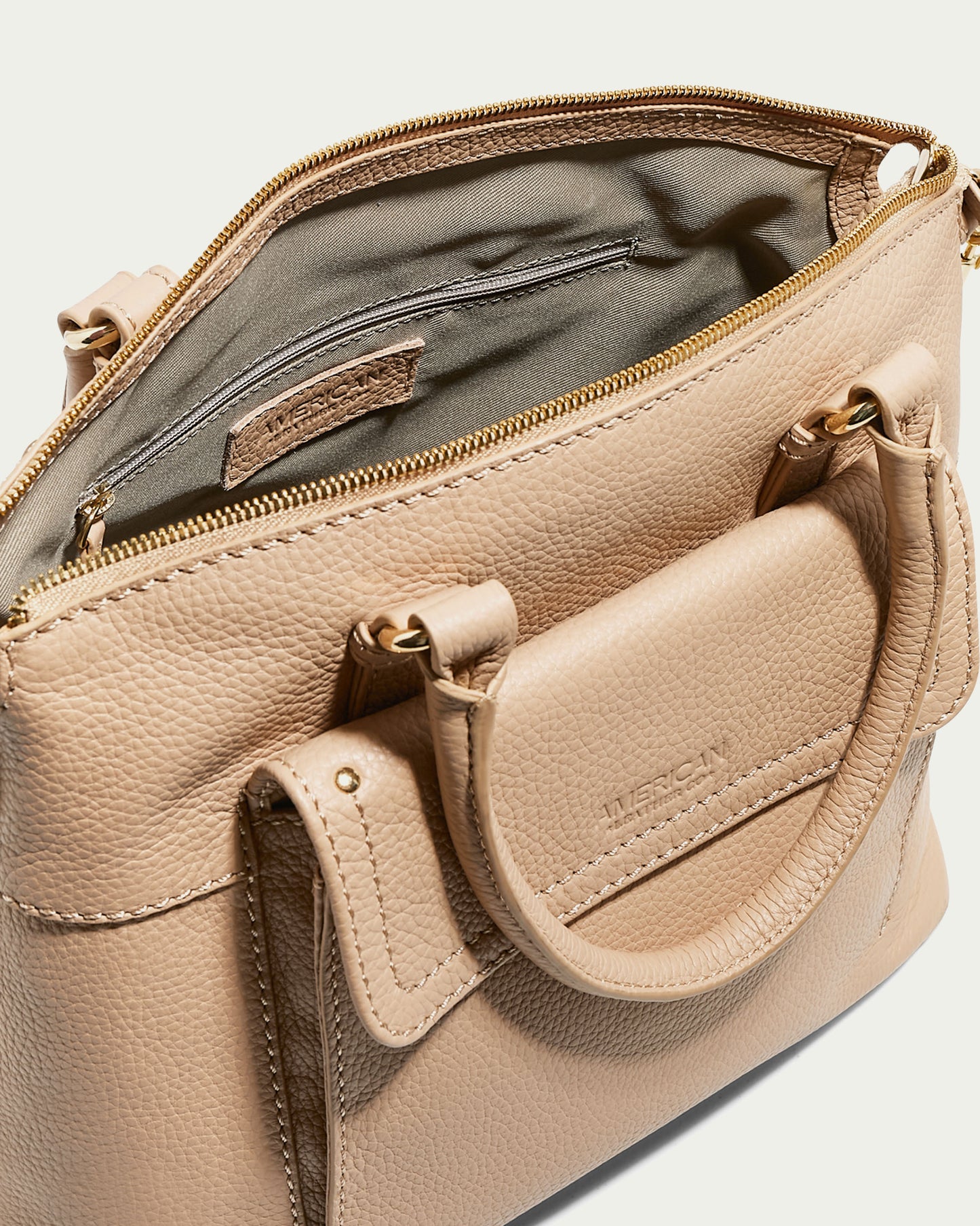 A beige Vona Convertible Dome Satchel handbag with a partially open top zipper reveals an interior with a zippered pocket. This elegant piece, made from genuine leather, features two handles and detailed stitching. The brand name "American Leather Co." is embossed on a small tag inside the bag and also on the exterior pocket flap.