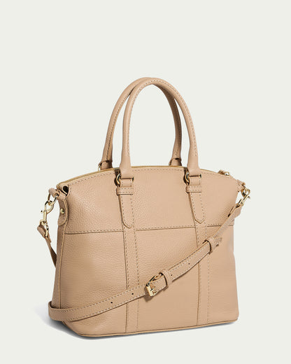 The Vona Convertible Dome Satchel by American Leather Co. is a beige genuine leather handbag with dual rolled handles and an adjustable, detachable shoulder strap. It features gold-tone hardware, including a clasp and buckles, and boasts a structured, trapezoidal shape with visible stitching details.