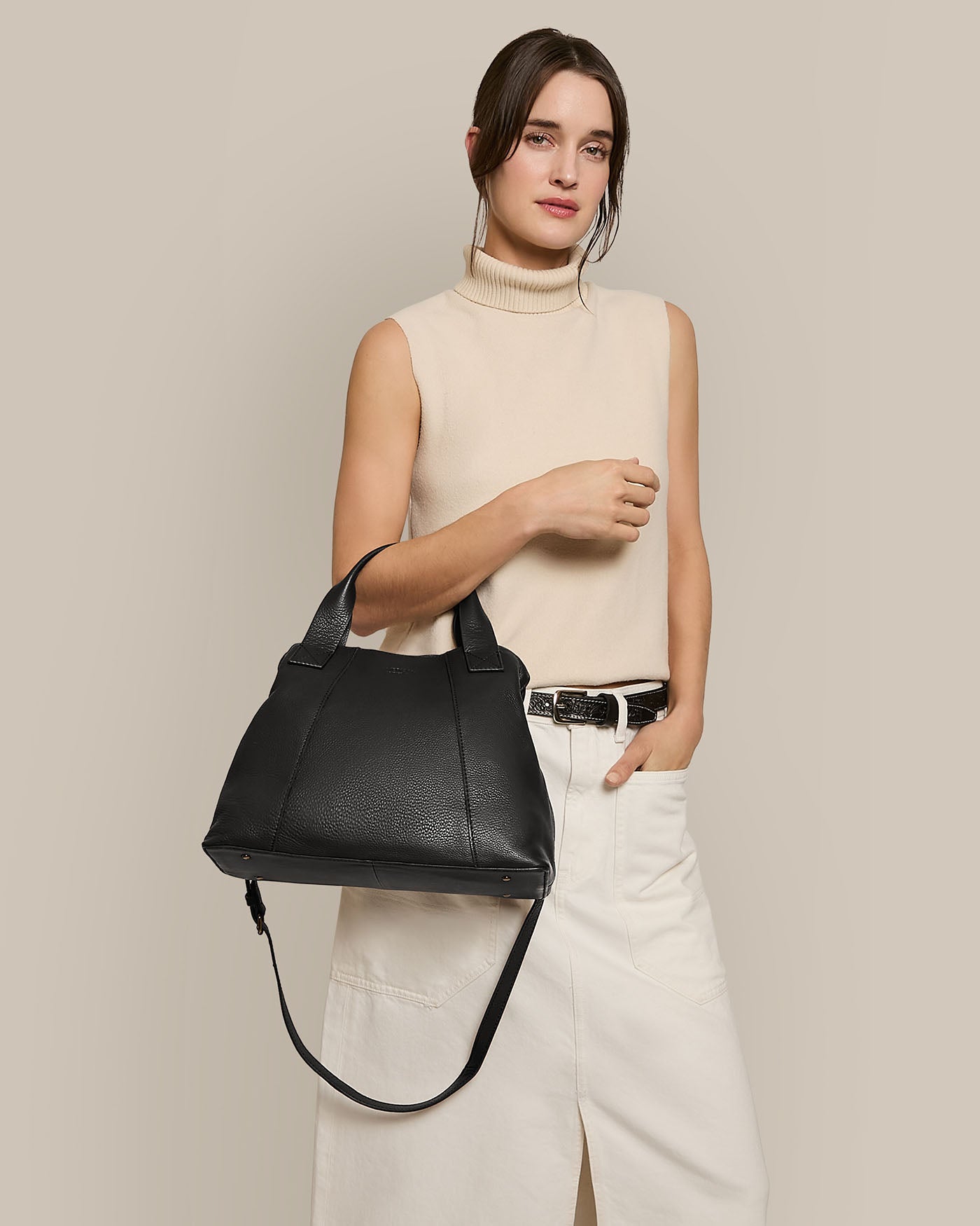 A woman elegantly poses with the Ada Triple Entry Satchel by American Leather Co., a structured black leather handbag with an adjustable crossbody strap. She wears a sleeveless beige turtleneck and white skirt against a plain beige background, highlighting the genuine American leather.