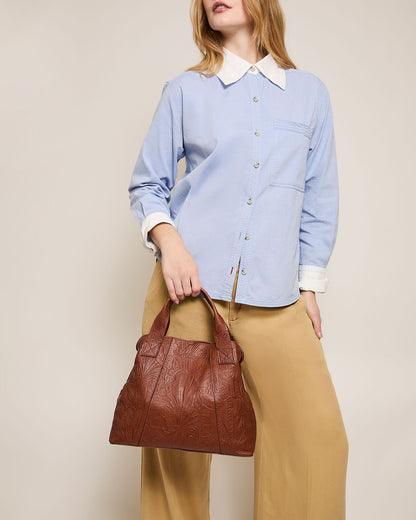 A woman in a light blue button-up shirt and beige pants holds the Ada Triple Entry Satchel by American Leather Co. With shoulder-length hair, she stands against a plain light background, her hand confidently resting on her hip.