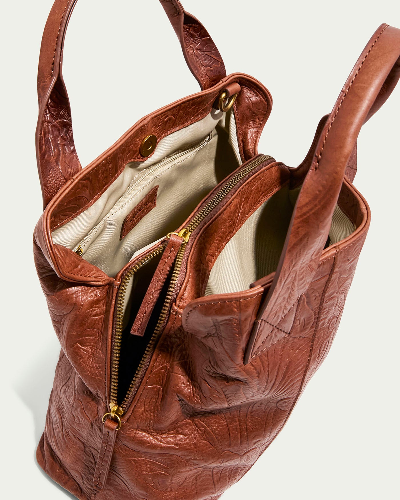 The Ada Triple Entry Satchel by American Leather Co. is a brown textured leather handbag with embossed floral patterns and two handles, showcasing a beige interior. It includes a central zippered compartment and a buttoned pocket for elegant organization.