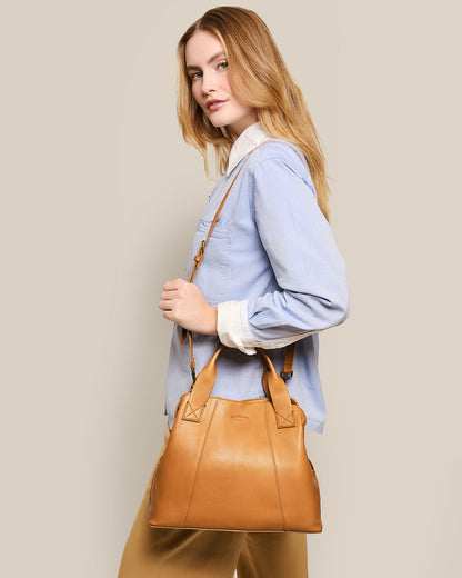 A person with long hair dons a light blue shirt and holds the Ada Triple Entry Satchel by American Leather Co., featuring an adjustable crossbody strap. The bag's genuine American leather contrasts beautifully against the plain beige background.