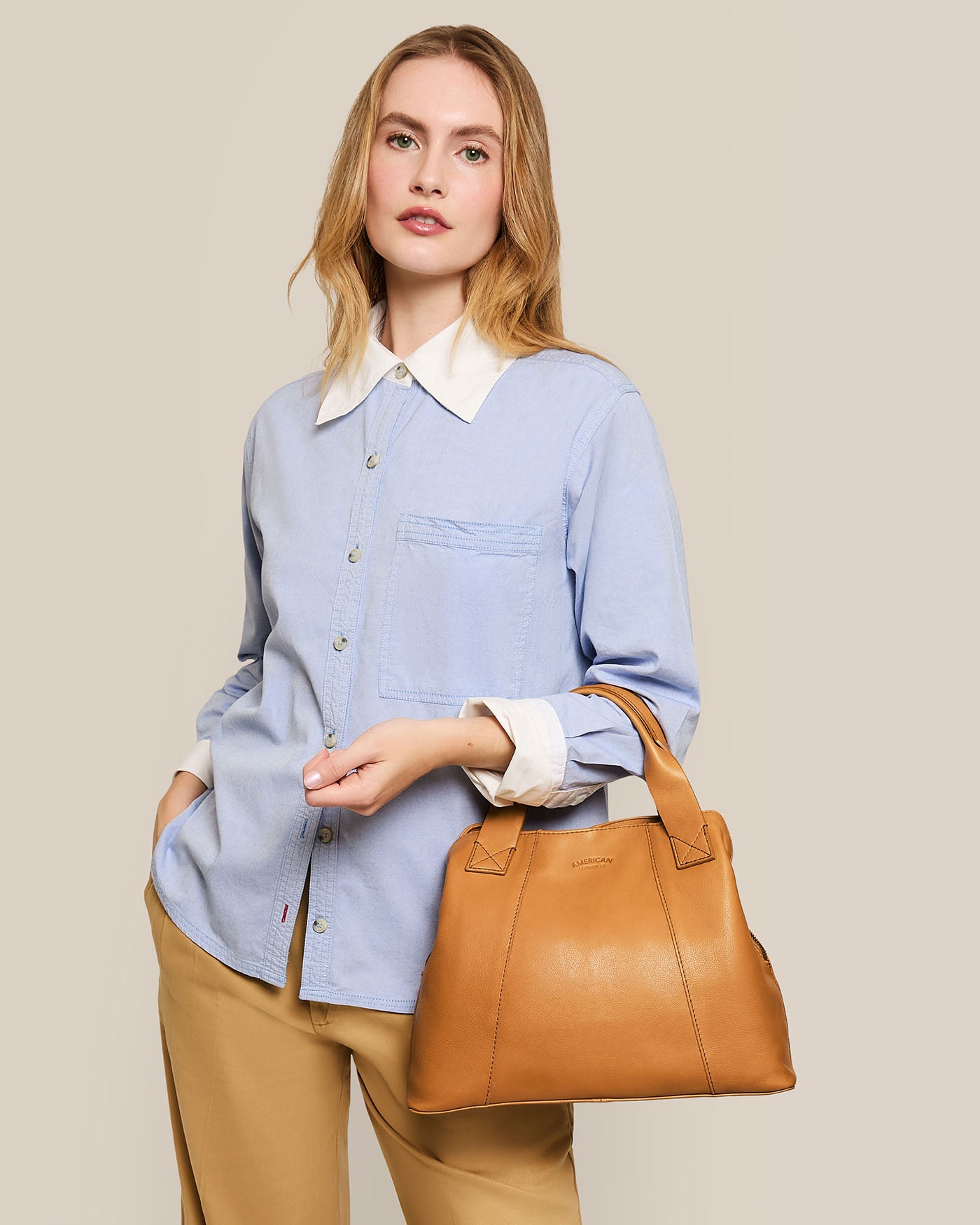 A person with long hair wearing a light blue button-up shirt and beige pants holds the Ada Triple Entry Satchel by American Leather Co. Against a beige background, they look forward, showing off the satchel's adjustable crossbody strap and genuine American leather craftsmanship.