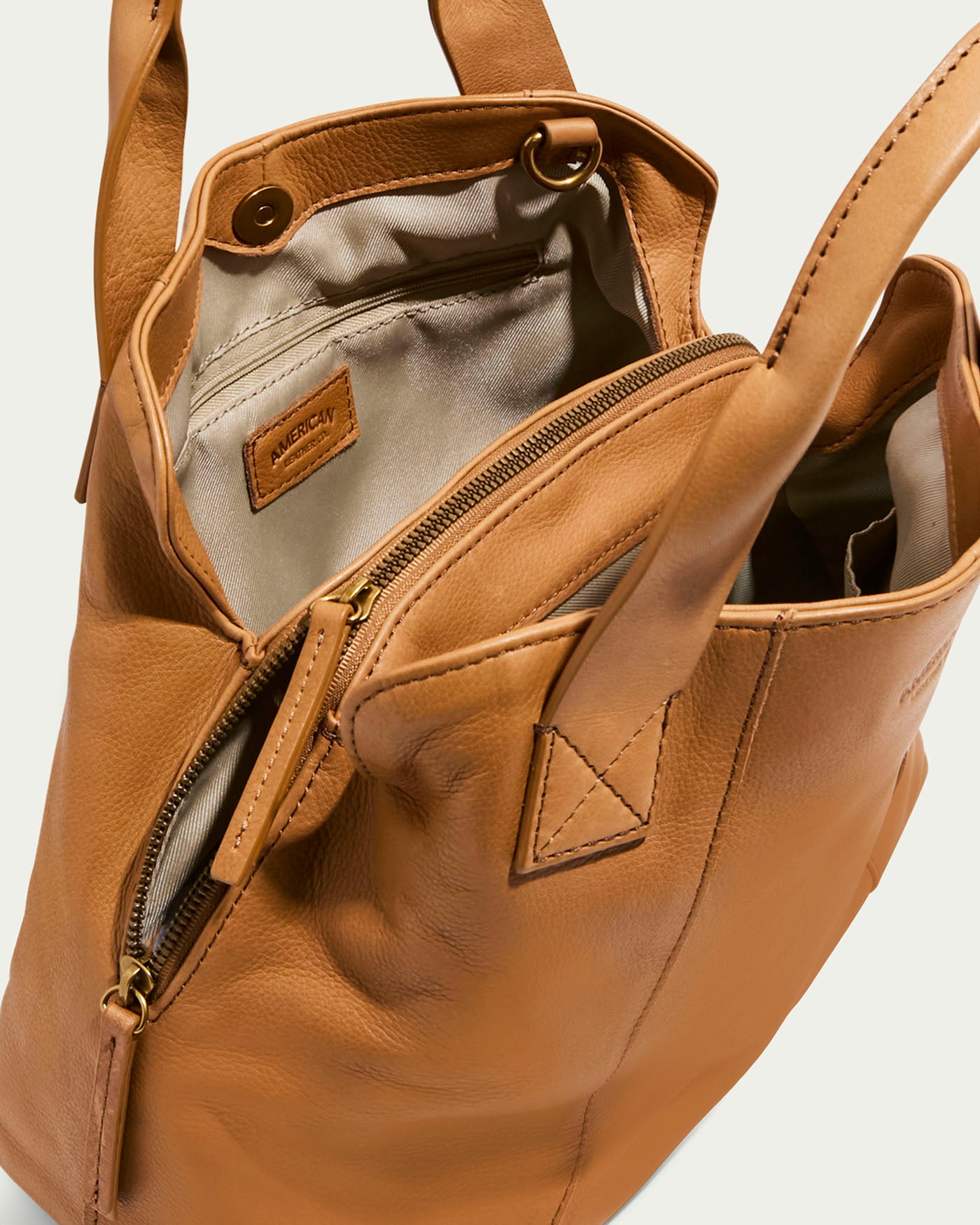 The Ada Triple Entry Satchel by American Leather Co. is a brown leather handbag with two handles and an adjustable strap, standing upright to show its beige interior lining with a zipper pocket and visible stitched leather tag, all set against a plain background.