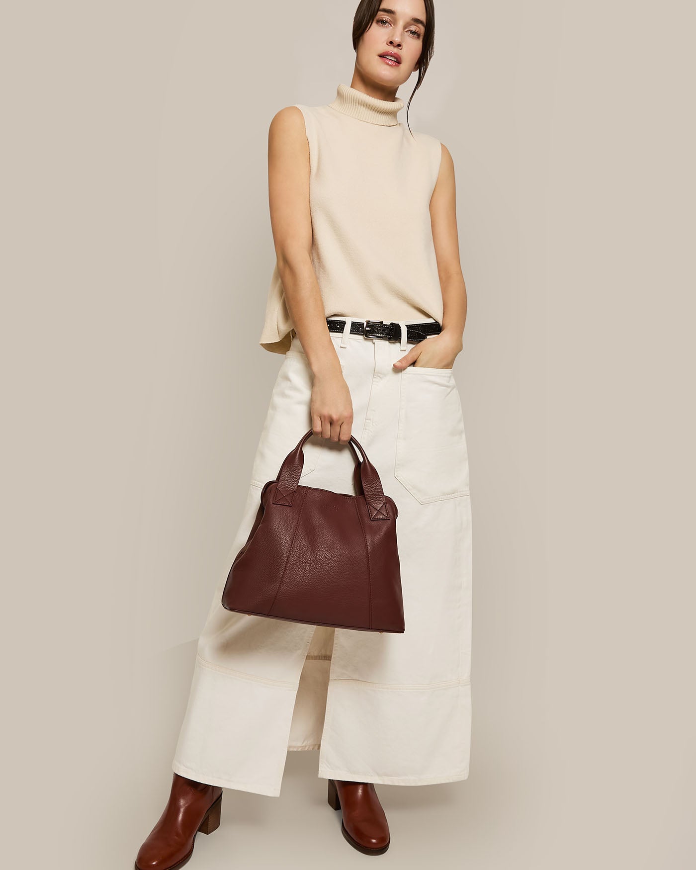 A person in a sleeveless beige turtleneck and wide-leg white pants carries the American Leather Co. Ada Triple Entry Satchel, a structured maroon bag with an adjustable crossbody strap. They pair it with brown ankle boots against a plain beige backdrop, creating a chic minimalist scene.