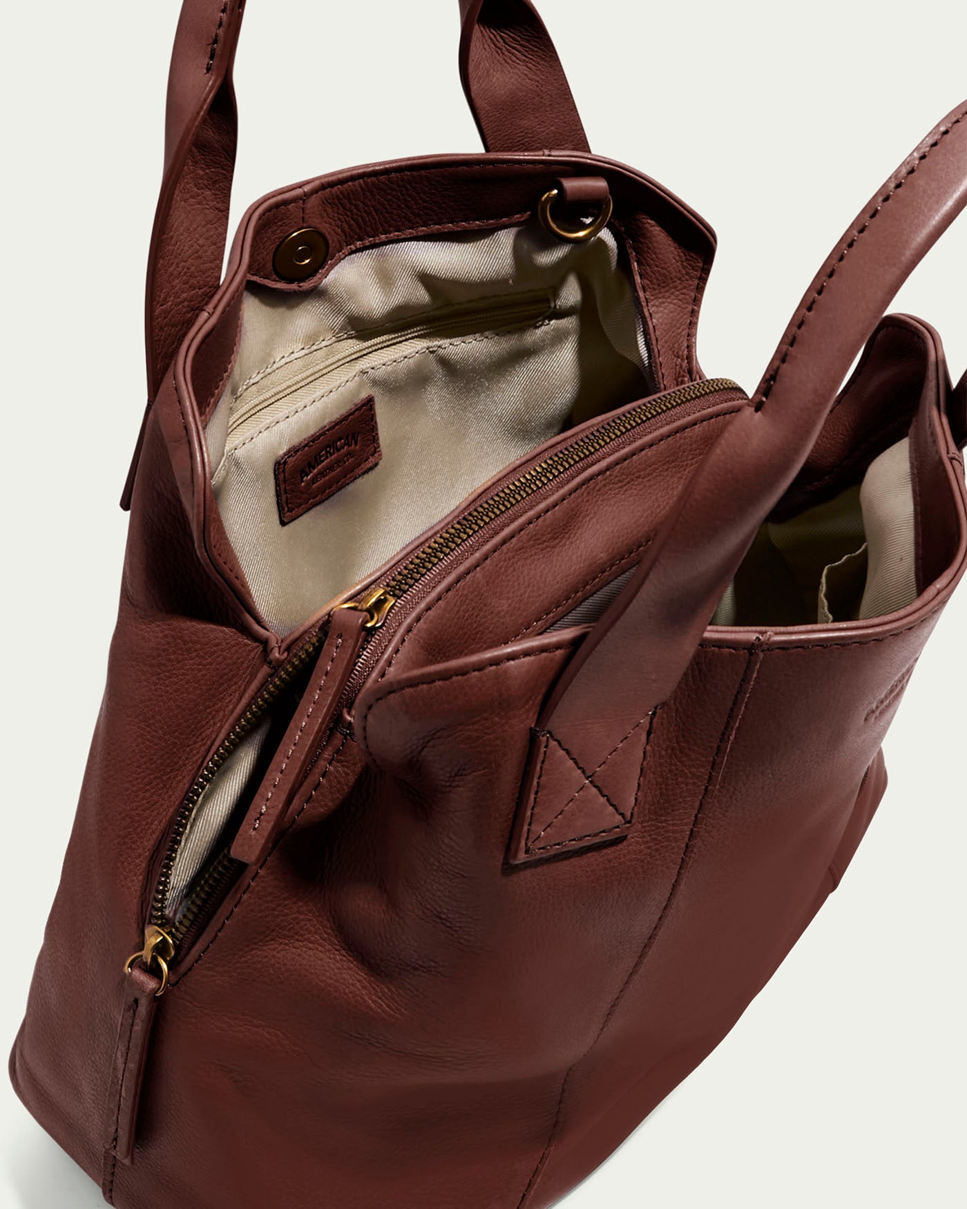 The Ada Triple Entry Satchel by American Leather Co. is a brown leather handbag featuring two handles, an adjustable crossbody strap, an open top showing its cream interior lining, a small inner pocket with a brand label, and a partially open outer zip pocket. Crafted from genuine American leather.