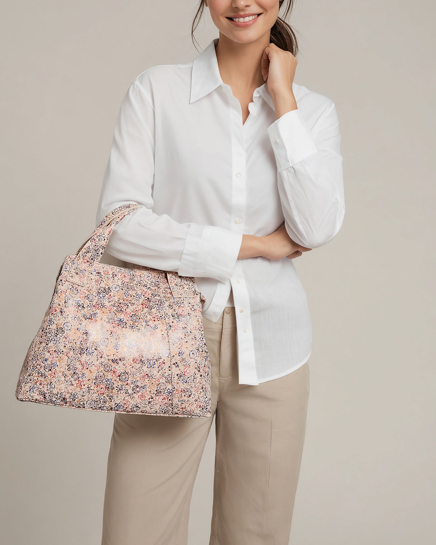 A person with tied-back hair, dressed in a white button-up and beige pants, holds the Ada Triple Entry Satchel by American Leather Co., featuring a floral pattern and adjustable crossbody strap. They stand against a plain, light backdrop, showcasing effortless style.