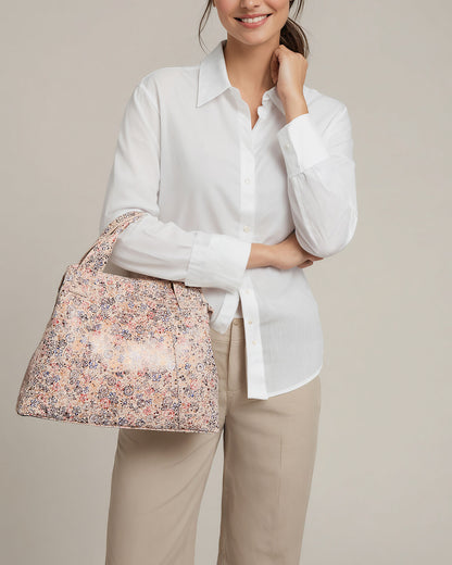 A person with tied-back hair, dressed in a white button-up and beige pants, holds the Ada Triple Entry Satchel by American Leather Co., featuring a floral pattern and adjustable crossbody strap. They stand against a plain, light backdrop, showcasing effortless style.