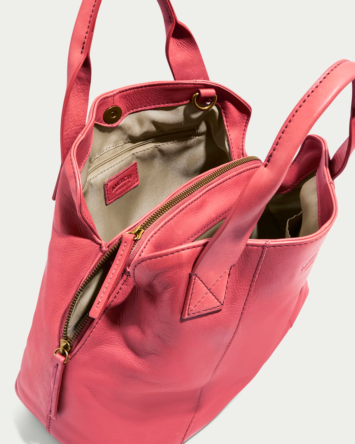 The Ada Triple Entry Satchel by American Leather Co. is a pink genuine leather tote with a structured design, featuring dual handles, a zipper closure, an open view of its beige interior, a small interior pocket, and a visible brand label.