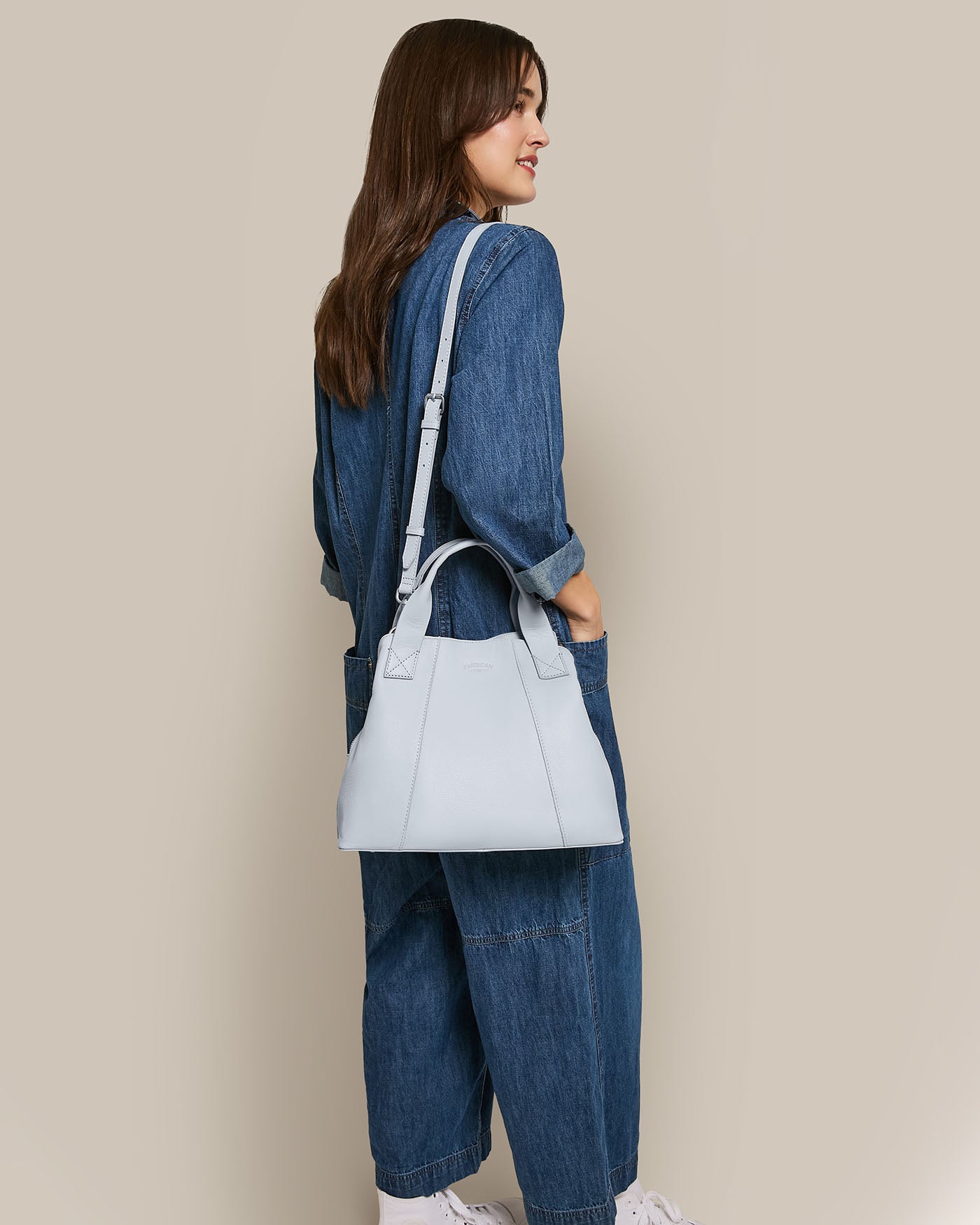 A person in a denim jumpsuit stands slightly sideways with long brown hair cascading down, carrying the American Leather Co. Ada Triple Entry Satchel over one shoulder against a plain beige background that highlights the scene's elegant simplicity.