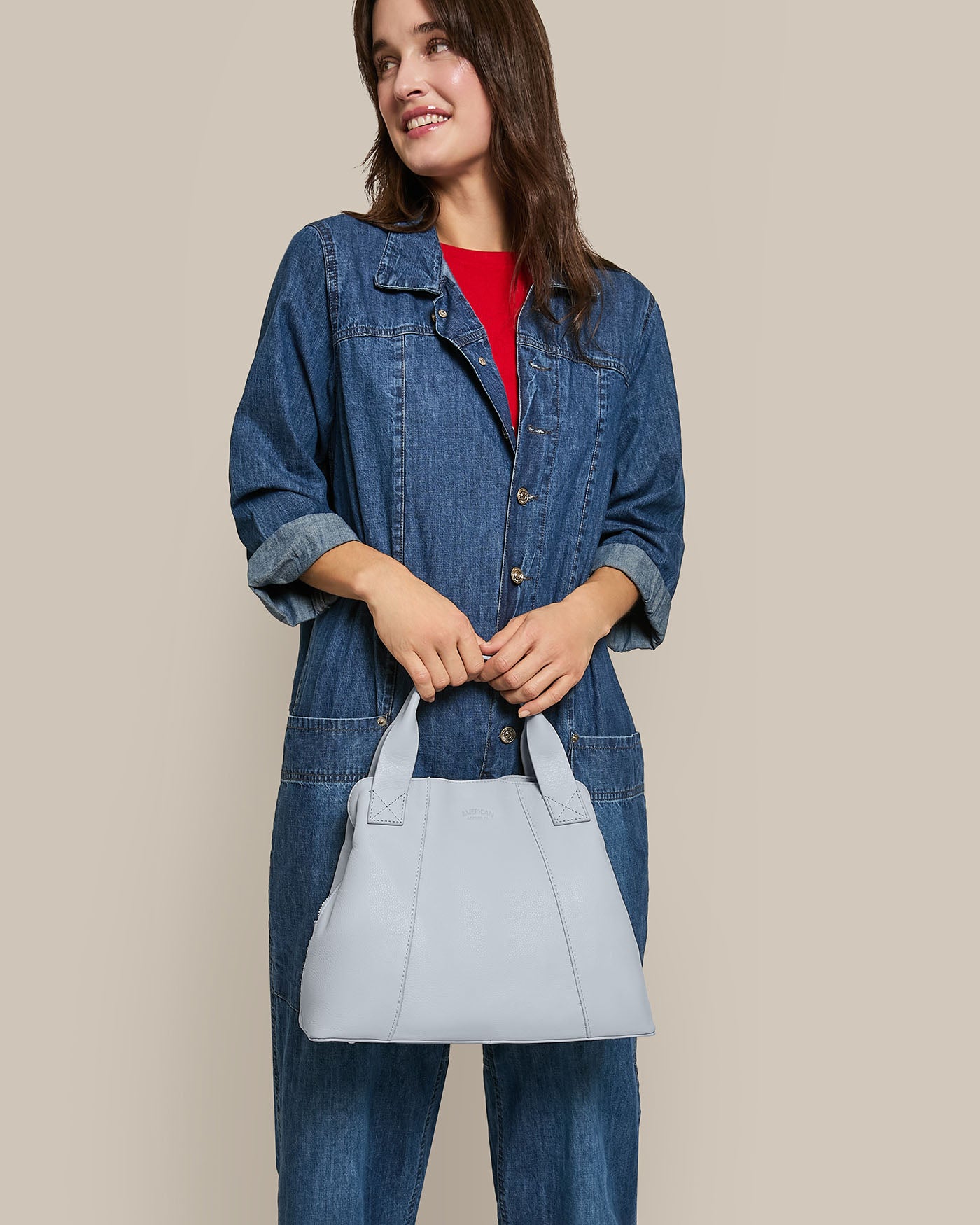 A woman with long hair smiles while holding the Ada Triple Entry Satchel from American Leather Co., made of genuine light gray American leather. She wears a denim jacket over a red shirt and stands against a plain beige background, exuding effortless style.