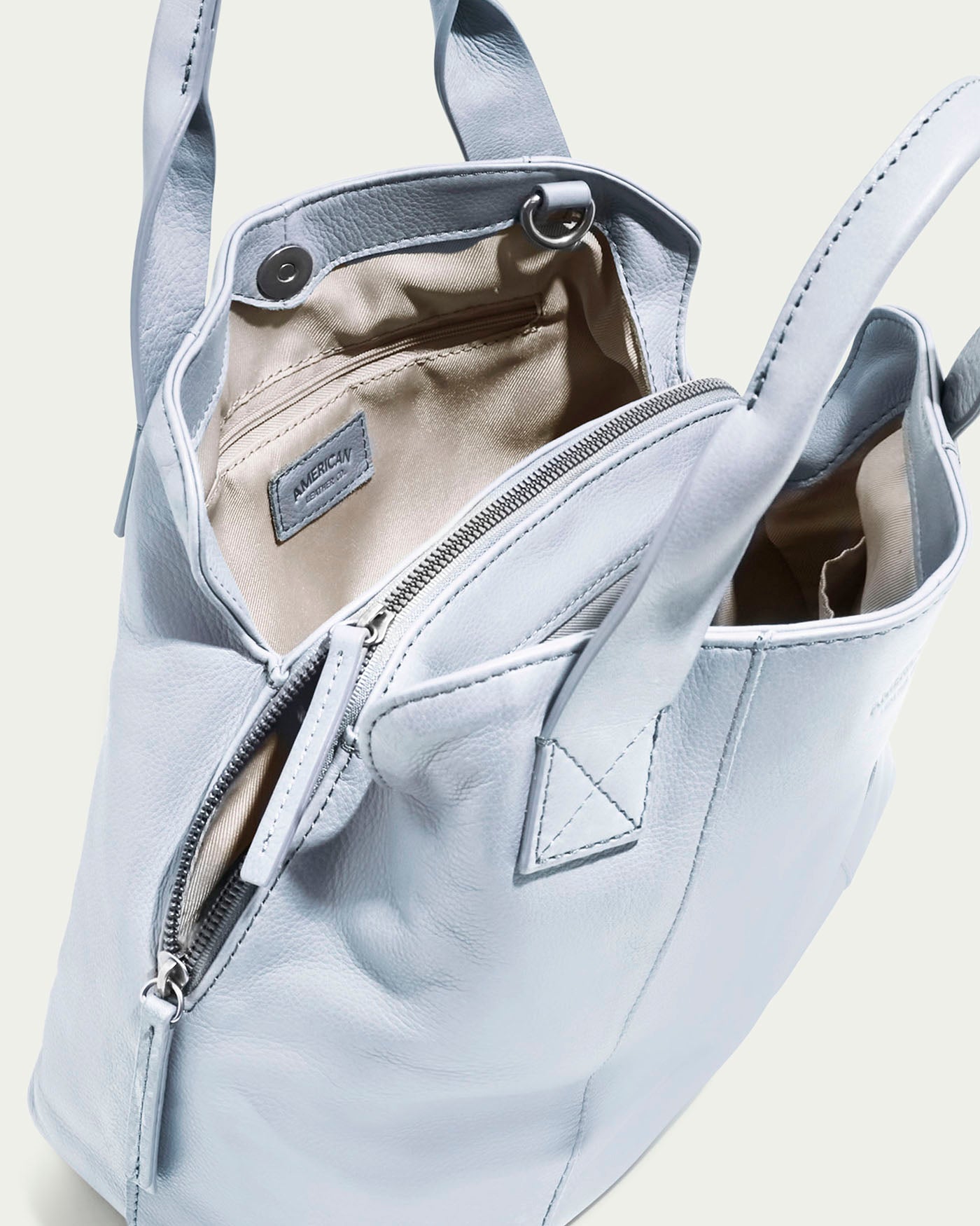 The Ada Triple Entry Satchel by American Leather Co. is a light blue leather tote with an open top and dual handles. Inside, it has a beige lining, zippered pocket, metal snap closure tab, and a charming small label near the pocket.