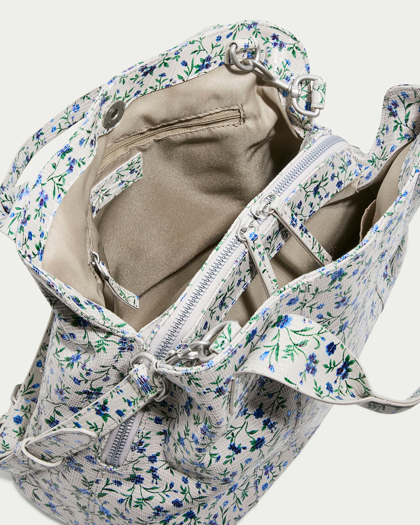 Top view of the Ada Triple Entry Satchel PLEASANTVILLE by American Leather Co. showcasing small blue and green flowers on white, with dual handles, an adjustable crossbody strap, and visible zippered compartment inside. Features include an attached side pocket and beige lining.
.