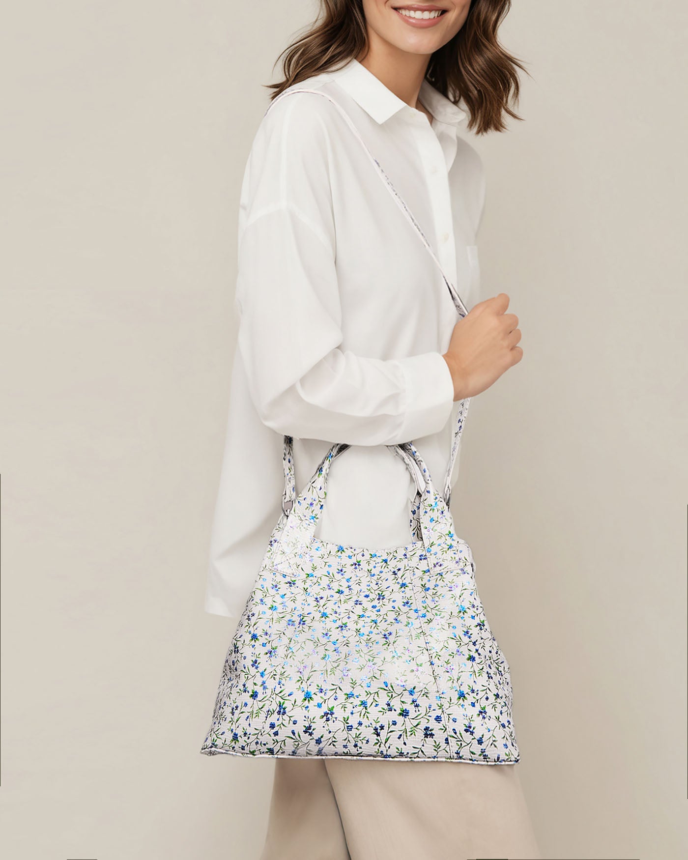 A person in a white shirt carries the Ada Triple Entry Satchel by American Leather Co., adorned with small blue and green flowers. Made from genuine American leather, it features handles and an adjustable crossbody strap. The plain background highlights this casual ensemble's subtle elegance.