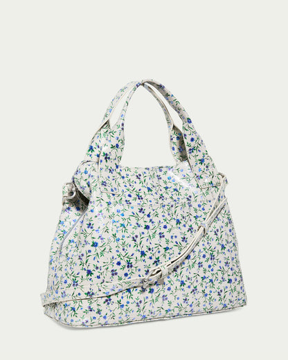 The Ada Triple Entry Satchel PLEASANTVILLE by American Leather Co. is a white handbag with a small blue and green floral pattern, crafted from Genuine American Leather. It features two short handles, an adjustable crossbody strap, and a structured shape for an elegant presentation.