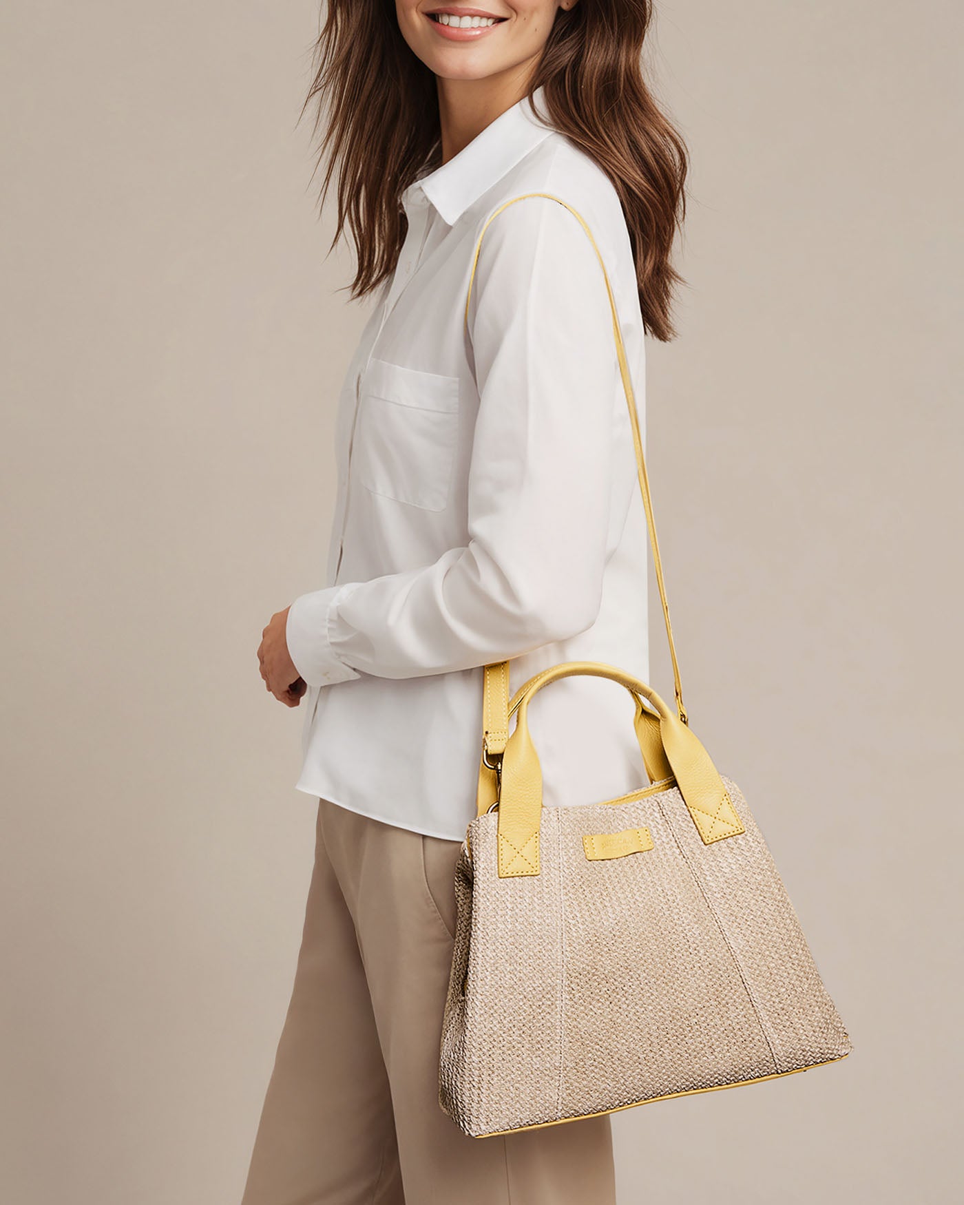 A person wearing a white shirt and beige pants carries the charming Ada Triple Entry Satchel by American Leather Co., featuring an adjustable crossbody strap with yellow accents. They are smiling and posing against a plain background.