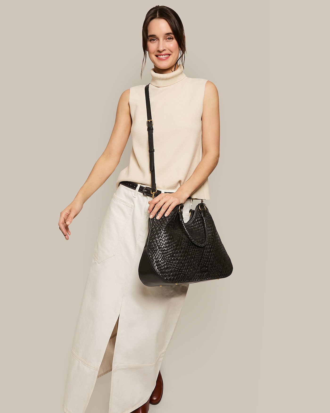 A woman wearing a sleeveless beige turtleneck and a long white skirt poses with a smile, carrying the Blair Convertible Tote by American Leather Co. over her shoulder. The plain beige background perfectly complements her stylish ensemble.