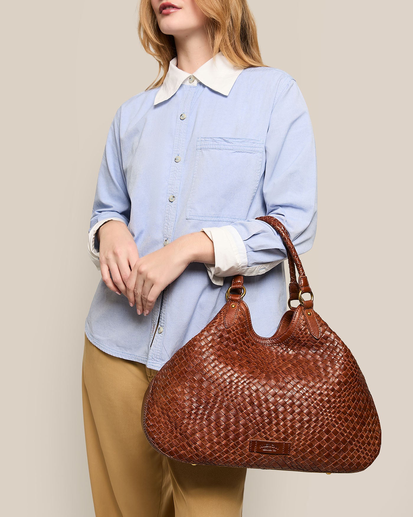 A person dons a light blue button-up shirt and beige pants while holding an American Leather Co. Blair Convertible Tote made from genuine woven leather. With the plain background and cropped head, the versatile bag stands out as the focal point.