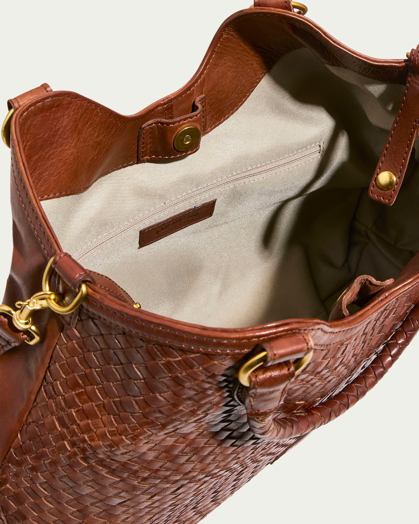 The Blair Convertible Tote by American Leather Co. is made from genuine woven leather with gold hardware. It features a light beige fabric lining, an open main compartment with a zippered pocket, decorative stitching, and a snap closure that enhance its versatile elegance.