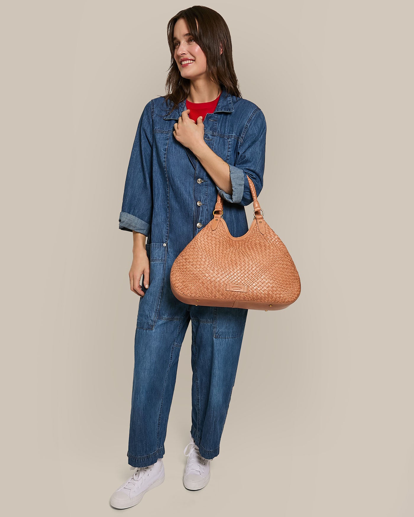 A person in a blue denim jumpsuit and white sneakers holds an American Leather Co. Blair Convertible Tote made of genuine woven leather, smiling and looking to the side against a plain background.