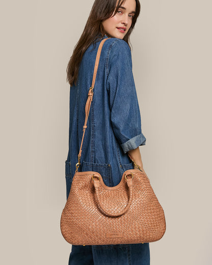 A person with long hair in a blue denim outfit is turned slightly to the side, showcasing an American Leather Co. Blair Convertible Tote made of genuine woven leather on their shoulder, standing out against the soft beige background.