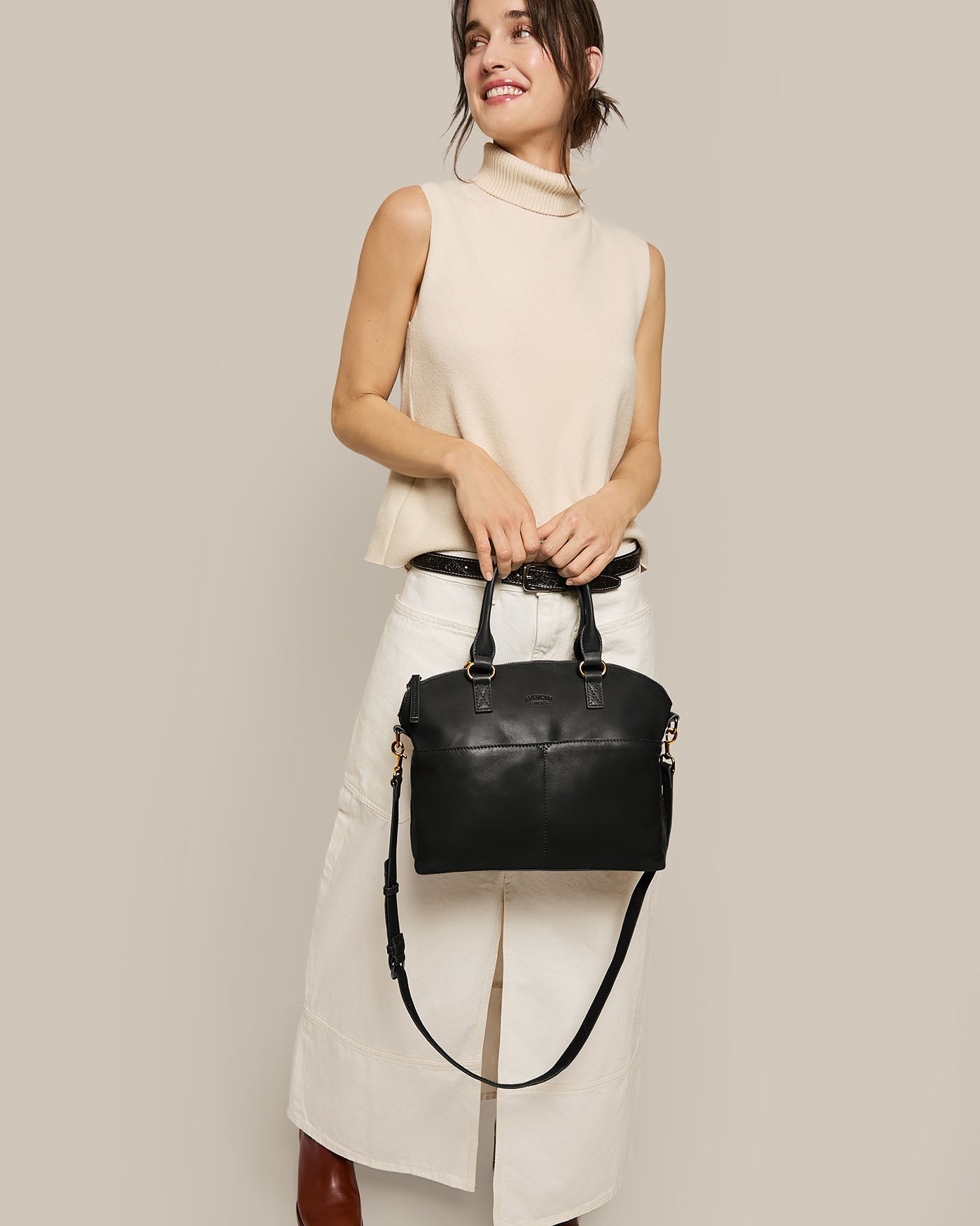 A woman in a sleeveless beige top and white skirt smiles with her hair tied back, holding the American Leather Co. Carrie Dome Satchel—a black leather bag with an adjustable crossbody strap—against a plain beige background.