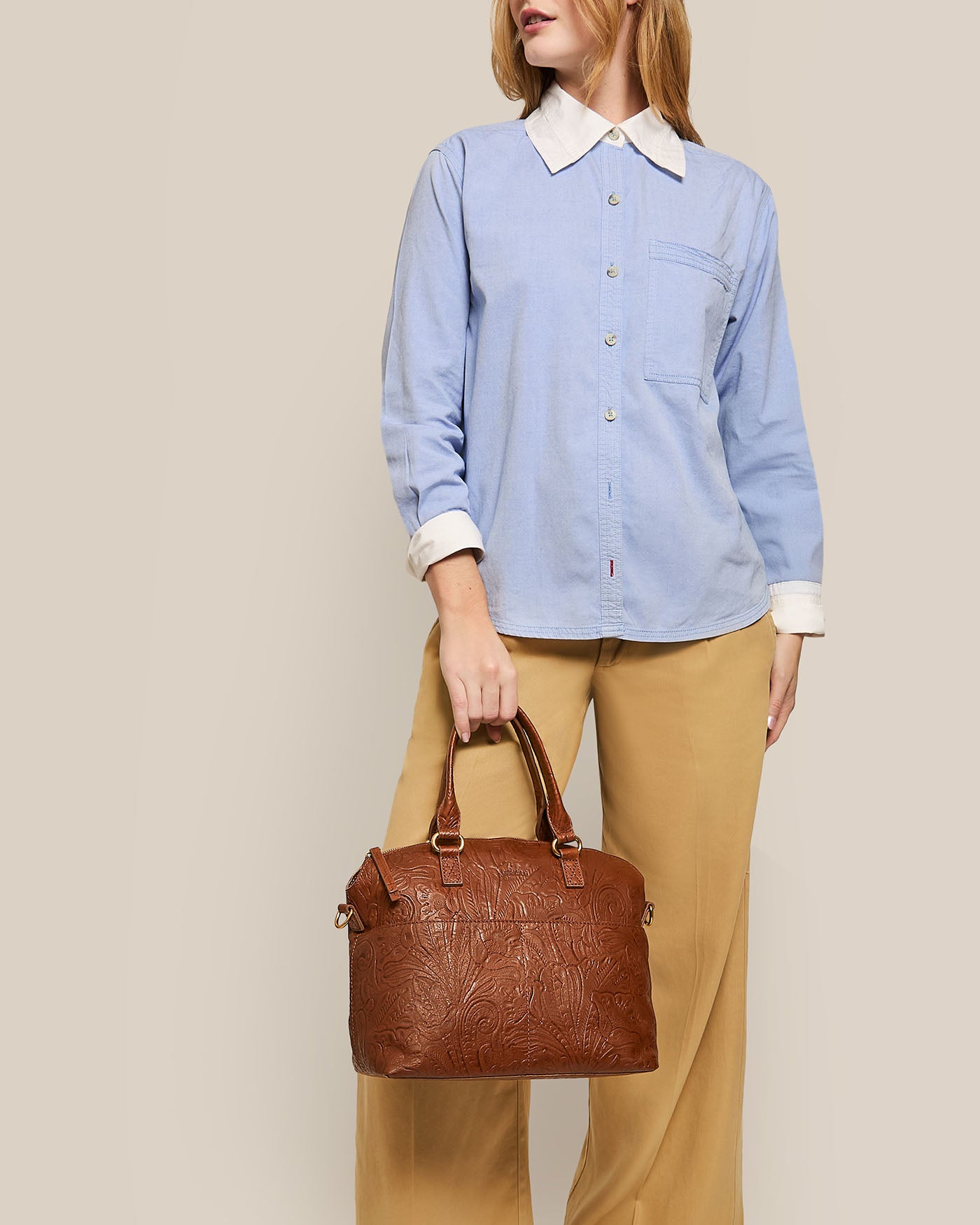 A person wearing a light blue button-up shirt with white collar and cuffs and beige wide-leg pants holds the Carrie Dome Satchel by American Leather Co., a brown embossed leather handbag with an adjustable crossbody strap, against a neutral background, showcasing their elegant style.