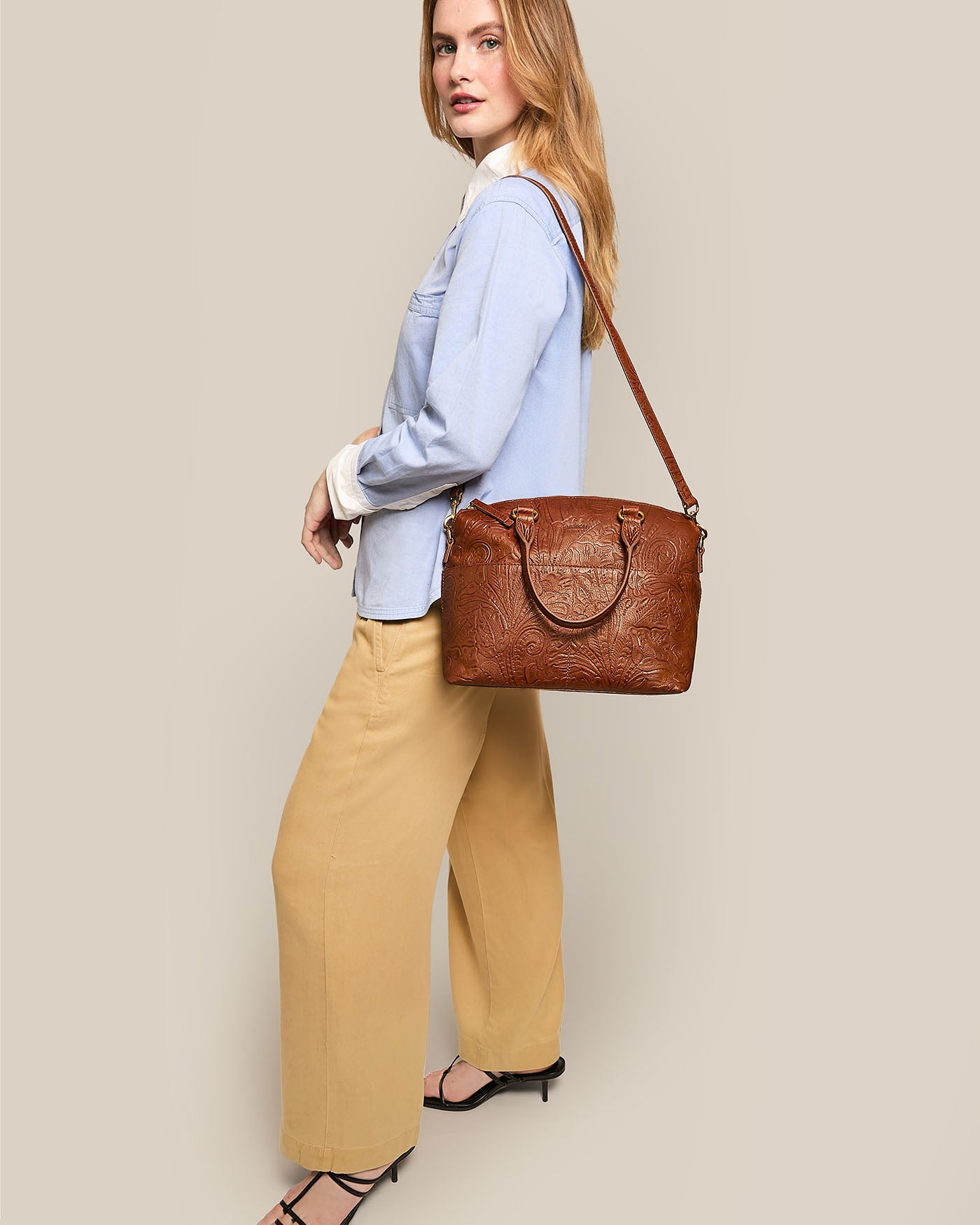 A woman with long hair stands sideways, wearing a light blue shirt and tan wide-leg pants. She holds a Carrie Dome Satchel by American Leather Co., crafted from genuine leather with embossed patterns, and wears black sandals against a neutral background.