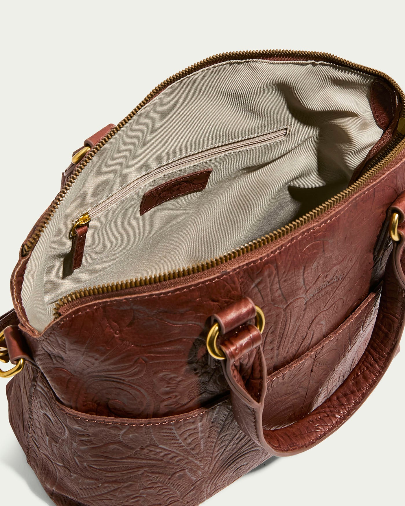 The Carrie Dome Satchel from American Leather Co. is a luxurious brown leather bag with embossed floral patterns. Crafted from genuine American leather, it features a beige fabric lining, a zippered pocket inside, two gold-ring handles, a top zipper closure, and an adjustable crossbody strap.