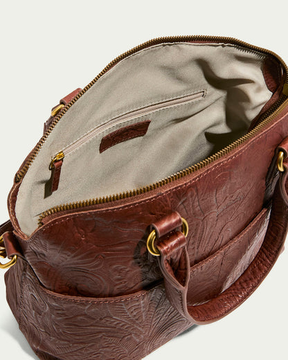 The Carrie Dome Satchel from American Leather Co. is a luxurious brown leather bag with embossed floral patterns. Crafted from genuine American leather, it features a beige fabric lining, a zippered pocket inside, two gold-ring handles, a top zipper closure, and an adjustable crossbody strap.