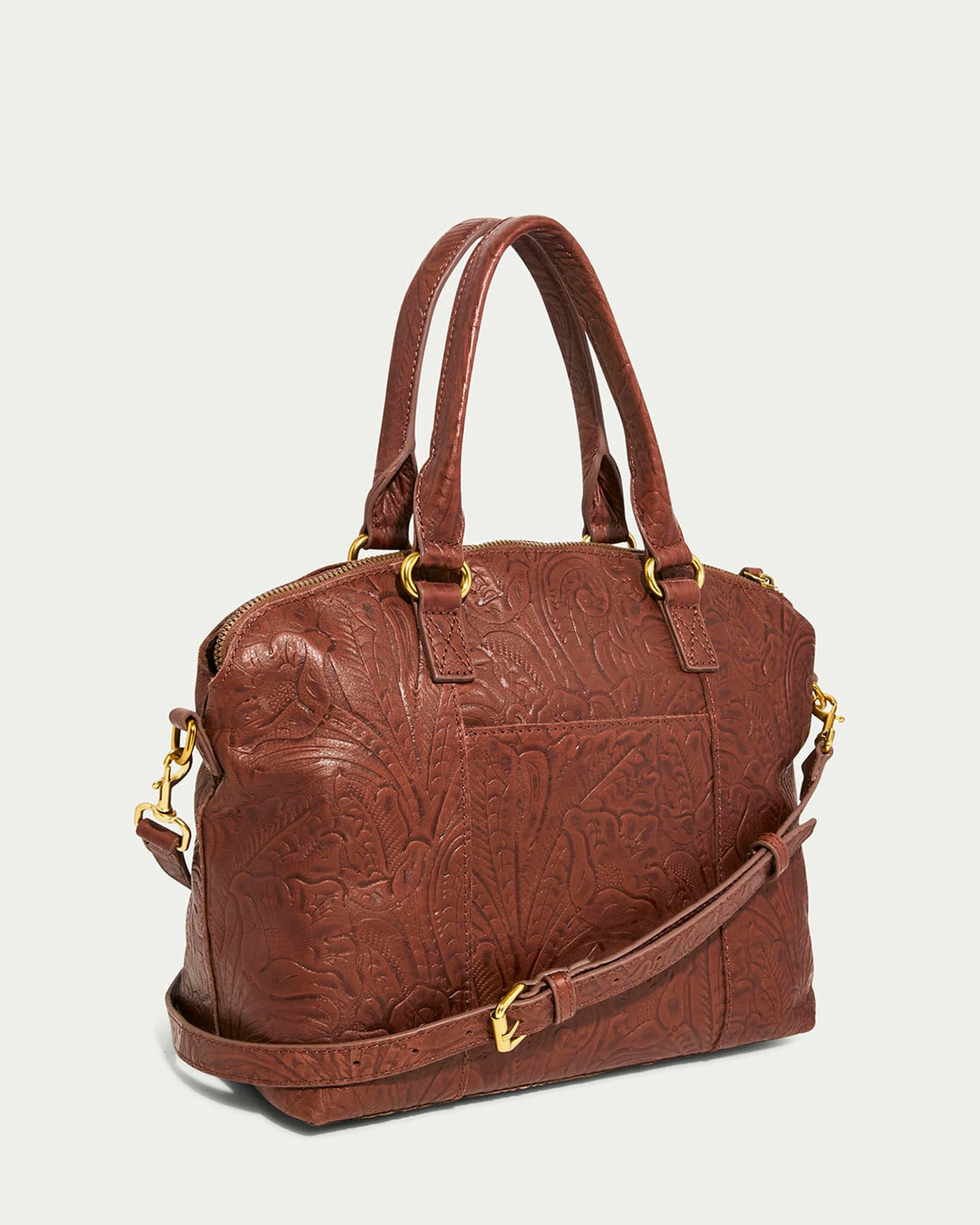 The American Leather Co. Carrie Dome Satchel is a brown leather handbag with an embossed floral design, dual top handles, an adjustable crossbody strap, and gold-tone hardware like a zipper closure and buckles, elegantly displayed on a light background.