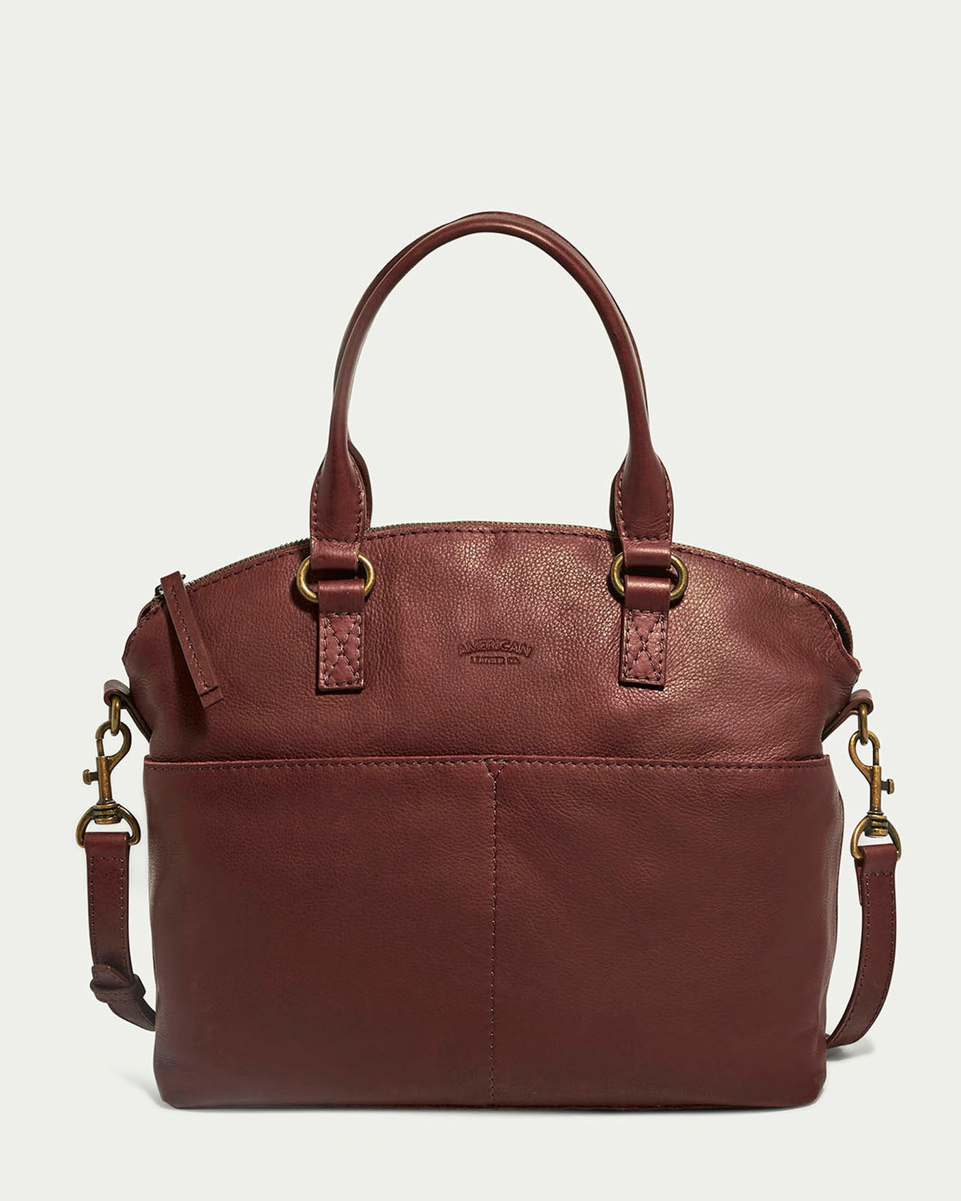 The Carrie Dome Satchel by American Leather Co. is a maroon leather bag with dual handles, an adjustable crossbody strap, brass hardware, and a top zipper closure. It boasts a sleek, rounded design with the brand logo subtly embossed on the front.