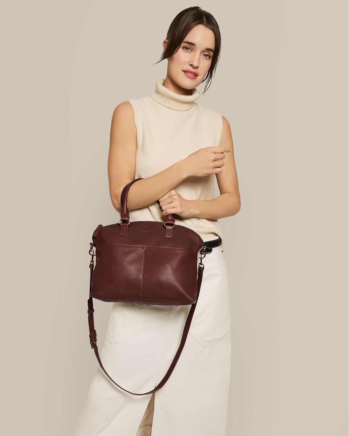 Against a plain beige backdrop, a woman with a slight smile holds the Carrie Dome Satchel, an American Leather Co. handbag in maroon. It features genuine leather and an adjustable crossbody strap, paired stylishly with her beige sleeveless turtleneck and white pants.