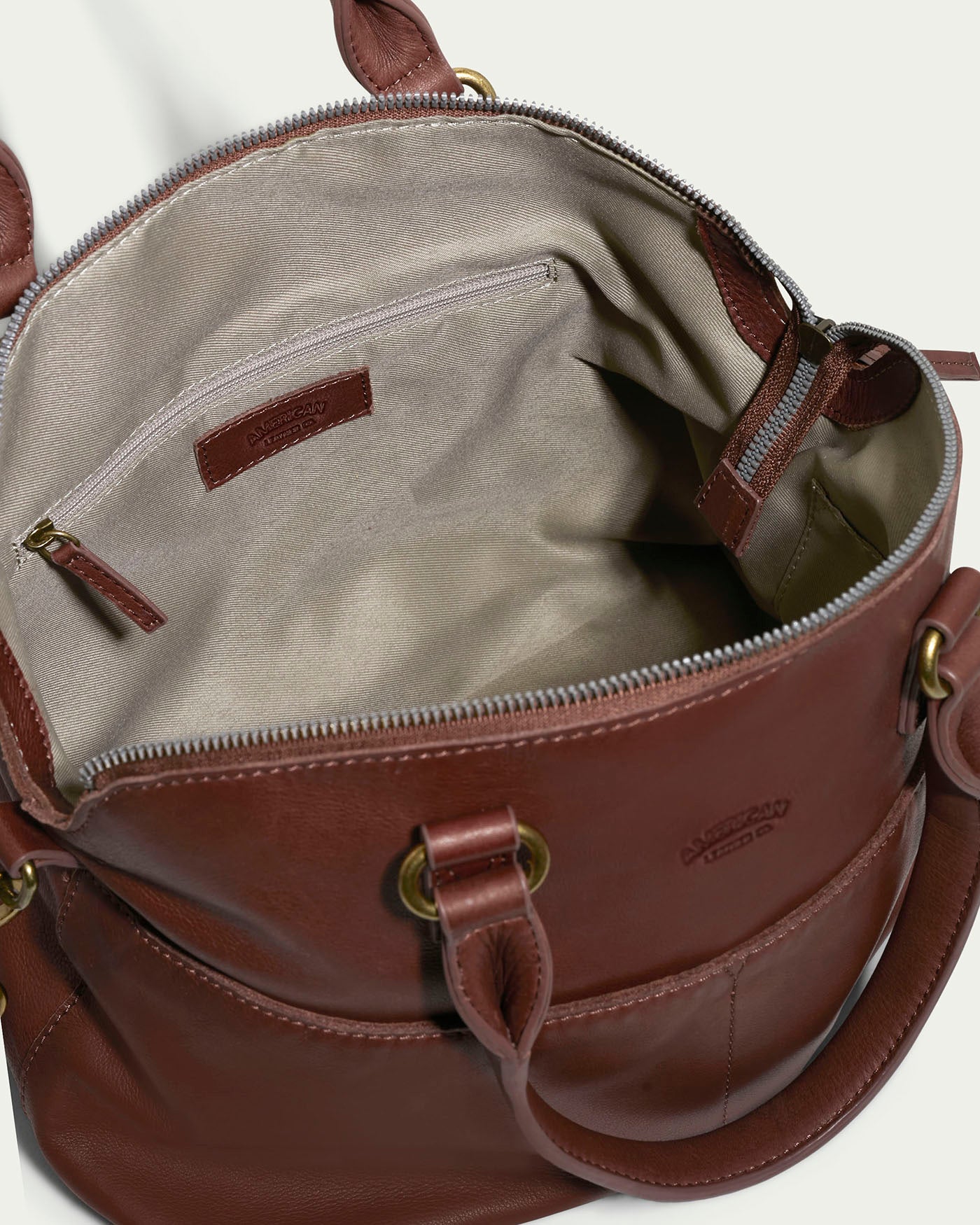 The Carrie Dome Satchel by American Leather Co. is an elegant leather bag crafted from genuine American leather, featuring a visible beige fabric interior with a zipper and open pocket. It includes metal hardware and an adjustable crossbody strap, with the brand name embossed on the leather.