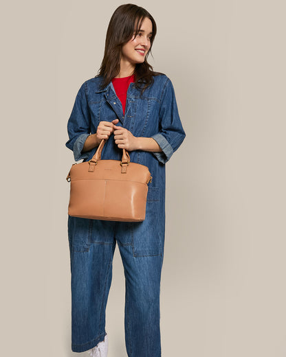 A person smiles in a denim jumpsuit and red shirt while holding the Carrie Dome Satchel by American Leather Co., featuring a tan leather body and crossbody strap, against a plain neutral background.