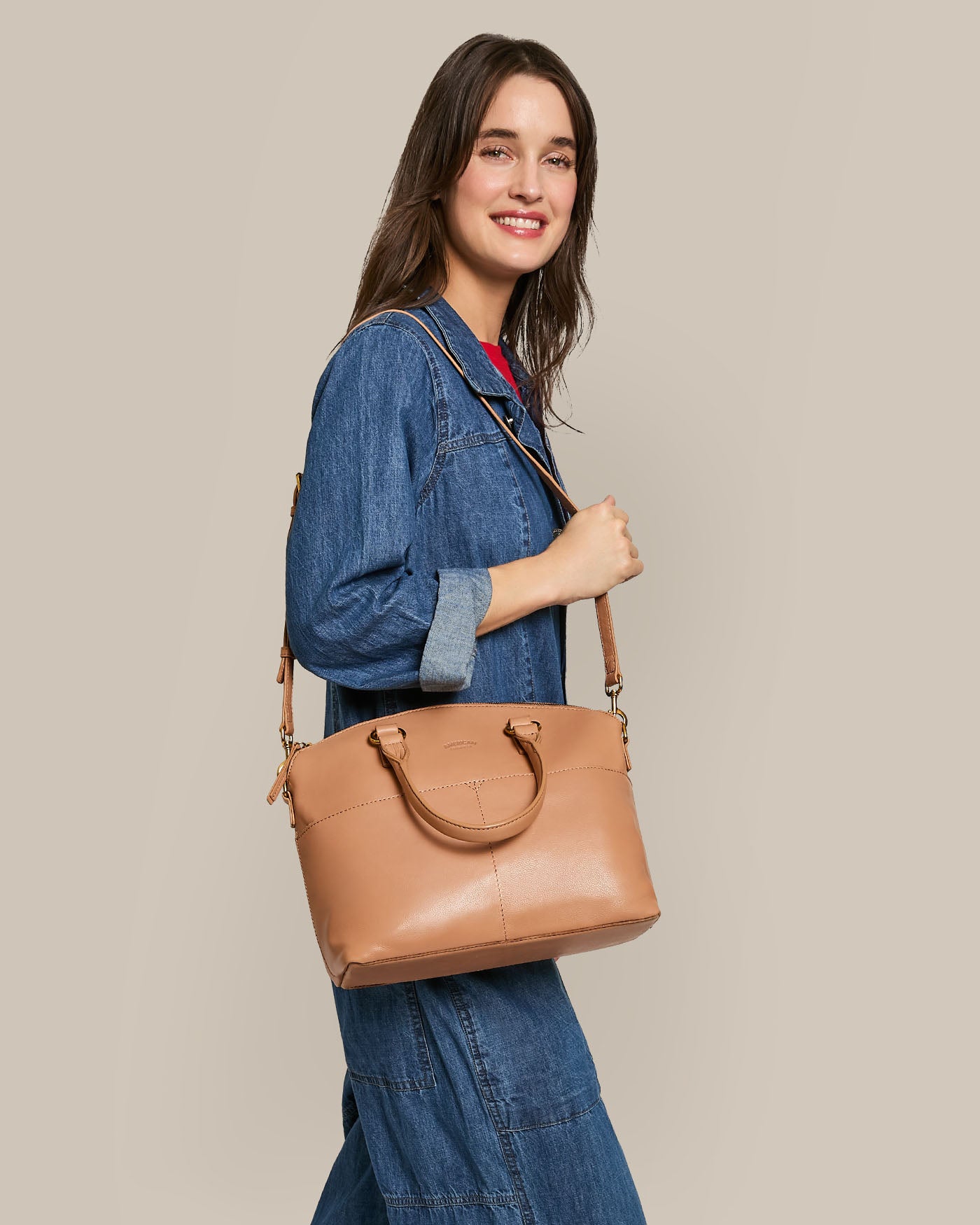 A denim-clad person smiles while holding the Carrie Dome Satchel by American Leather Co., crafted from genuine tan leather with a crossbody strap and handles, set against a plain beige background.