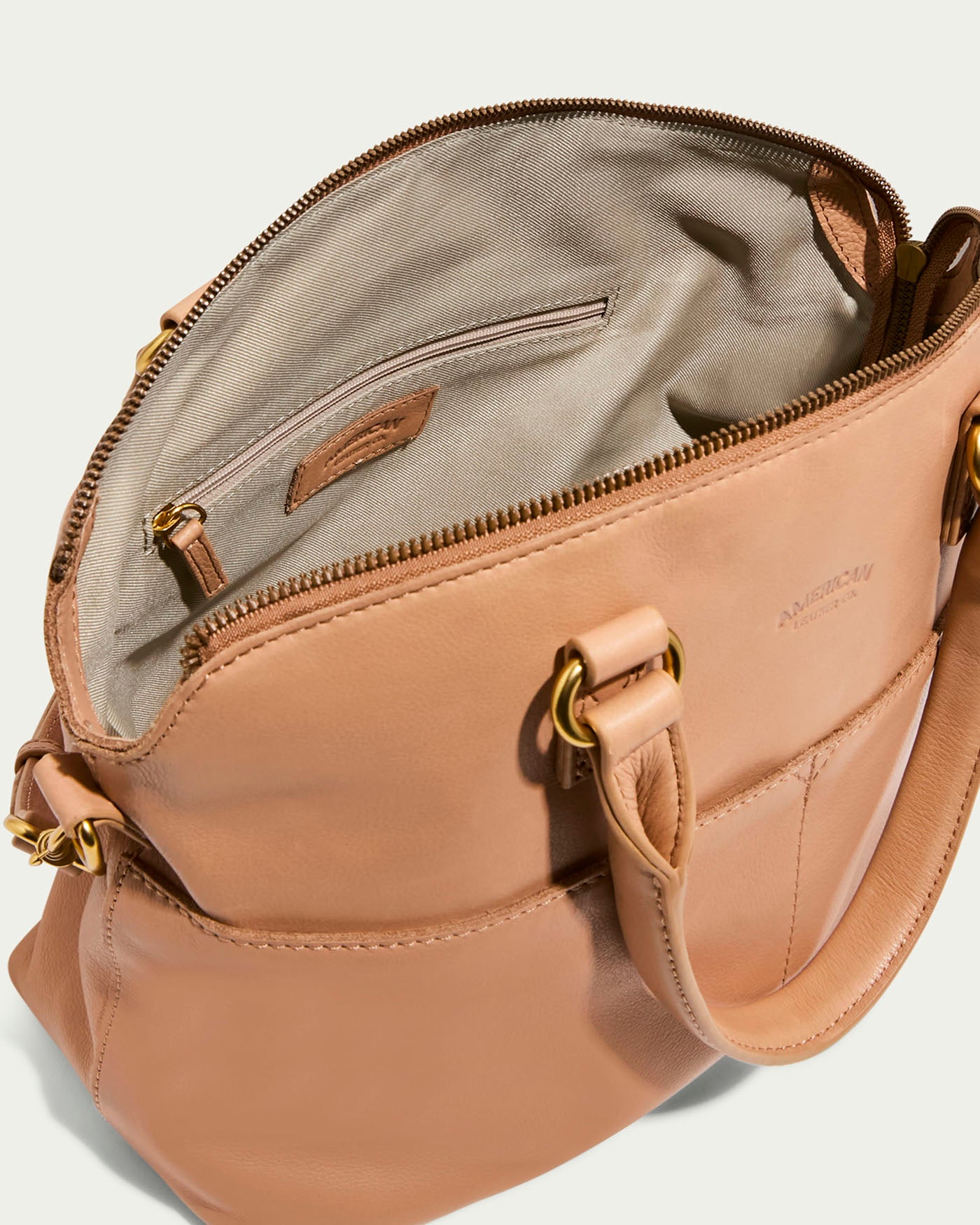 The Carrie Dome Satchel by American Leather Co. features gold hardware, a beige fabric lining with an interior pocket and leather tag. It boasts sturdy handles, a versatile crossbody strap, and an embossed brand logo on the exterior for added style.