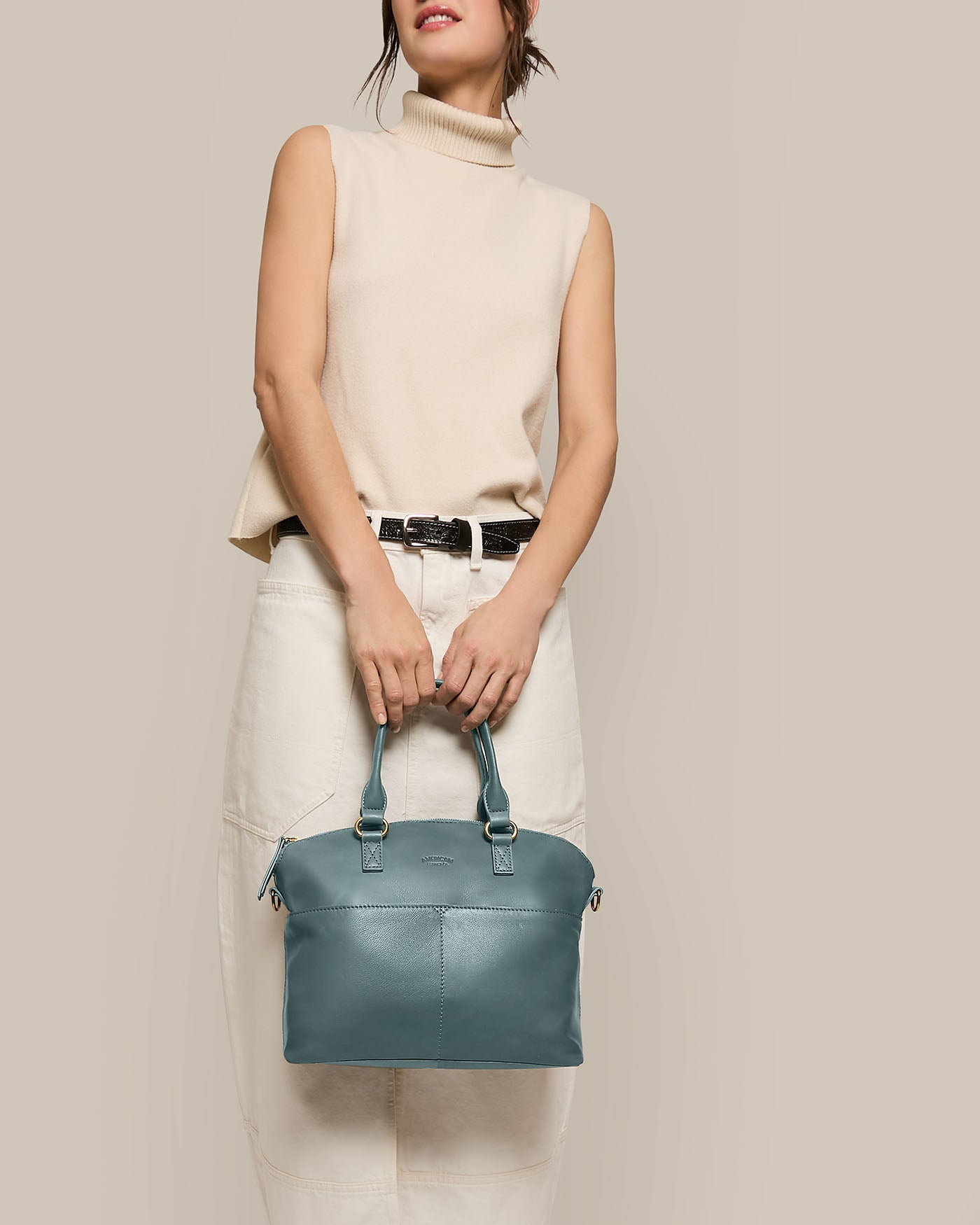 A person in a sleeveless beige turtleneck top and white skirt holds an American Leather Co. Carrie Dome Satchel, a teal handbag with an adjustable crossbody strap. Set against a soft beige background, the Genuine American Leather accessory is highlighted while the person's face remains unseen.