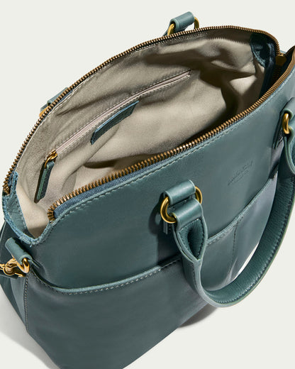 The Carrie Dome Satchel by American Leather Co. is an open teal leather handbag with gold hardware crafted from genuine American leather. It includes a spacious beige-lined interior with a zippered pocket, open compartments, zipper closure, adjustable crossbody strap, and two handles.