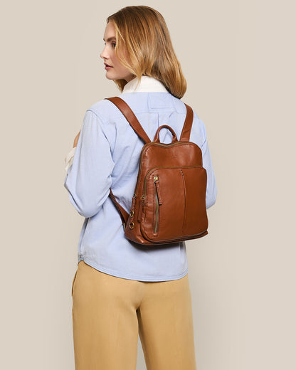 A woman with long hair, in a light blue button-up shirt and beige pants, faces slightly to the side while carrying the Cleveland Backpack by American Leather Co. This brown backpack purse, made of genuine leather, features multiple zippered compartments and adjustable straps against a neutral background.