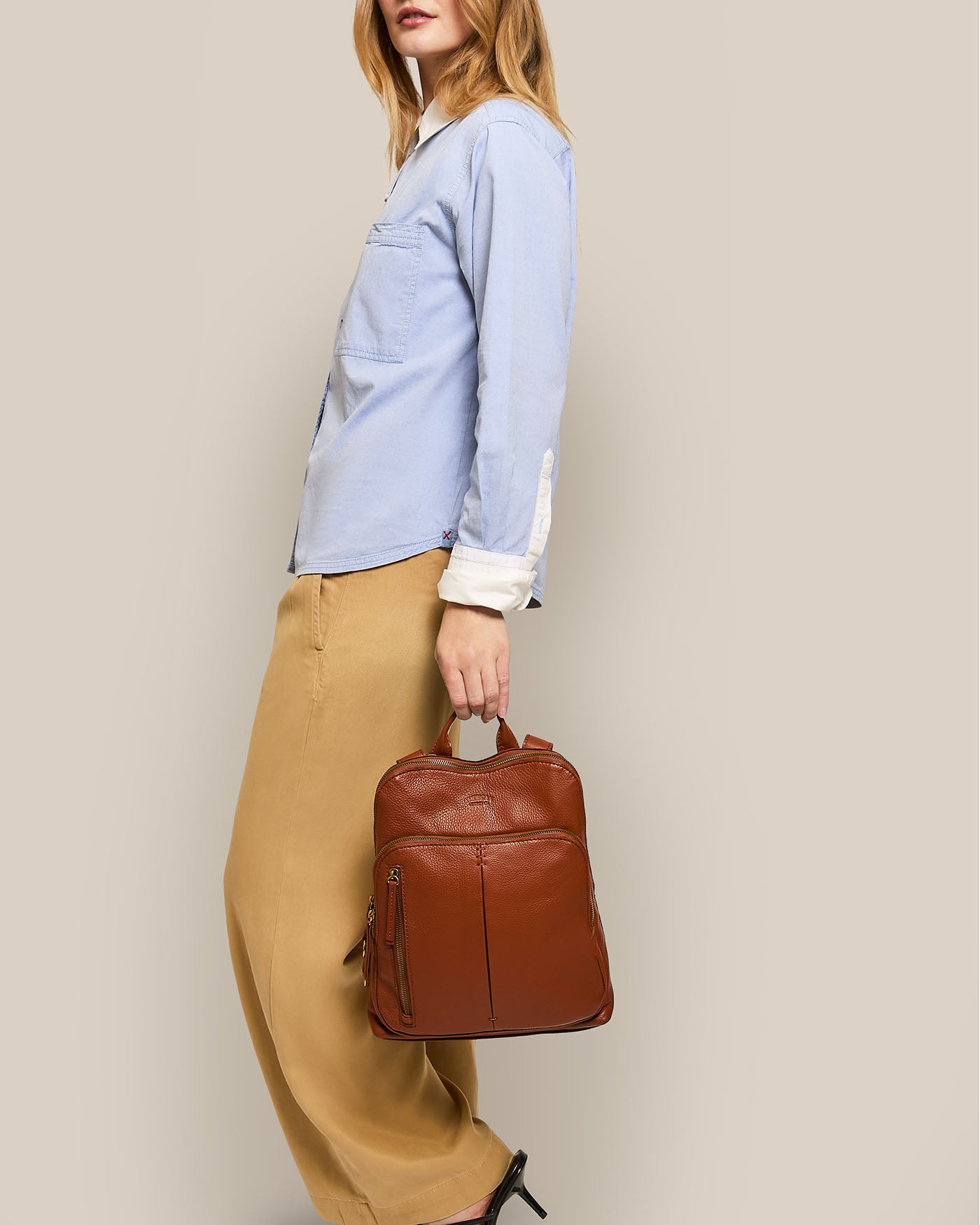 A person stands in profile against a neutral backdrop wearing a light blue button-up shirt and beige trousers. They hold the Cleveland Backpack by its top handle, showcasing its adjustable back straps. The American Leather Co. design keeps the person’s face out of view.