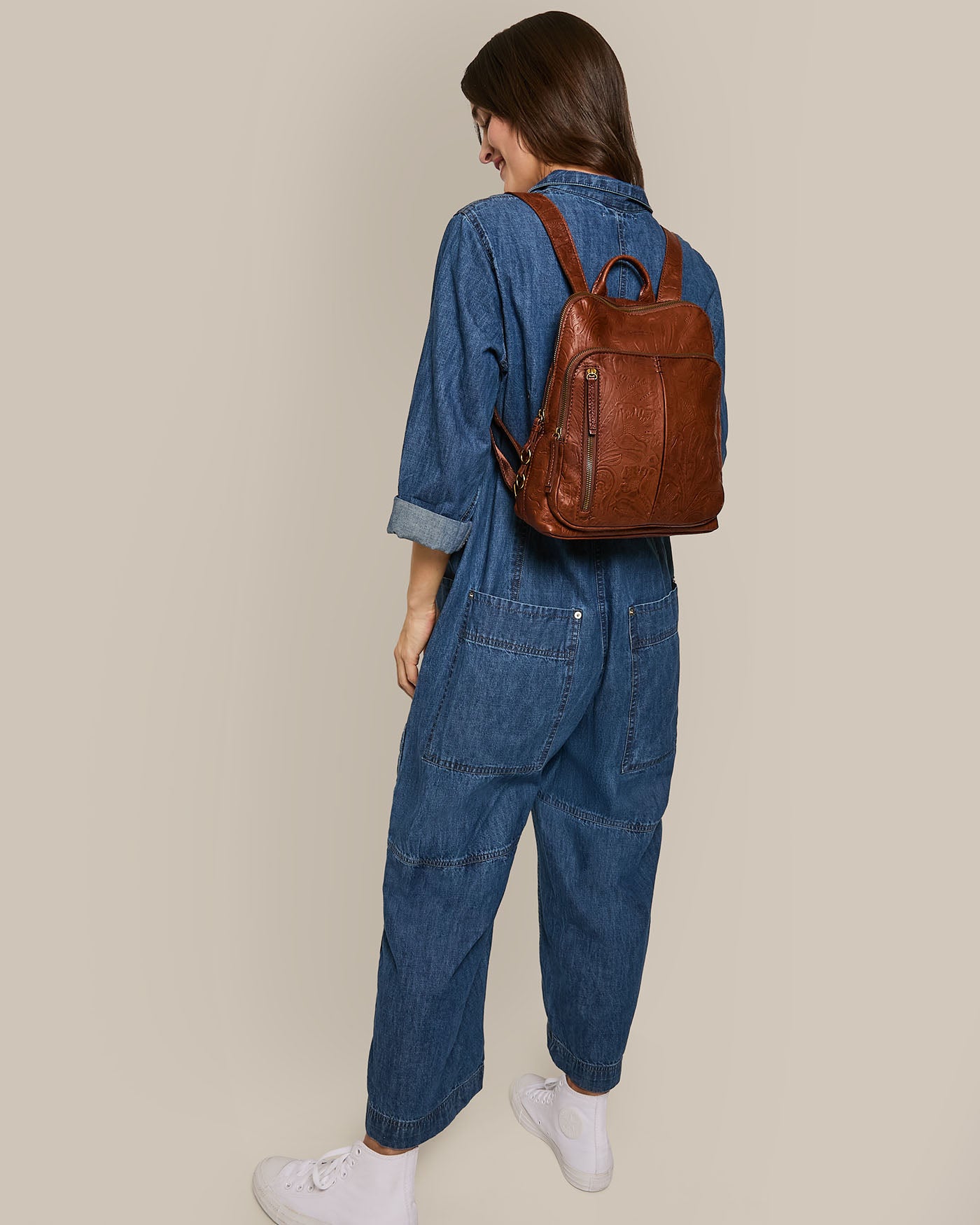 The woman, clad in a denim jumpsuit and white sneakers, glances down and to the side. Her Cleveland Backpack from American Leather Co., featuring antiqued hardware, stands out against the plain beige background, adding a touch of elegance to her casual ensemble.