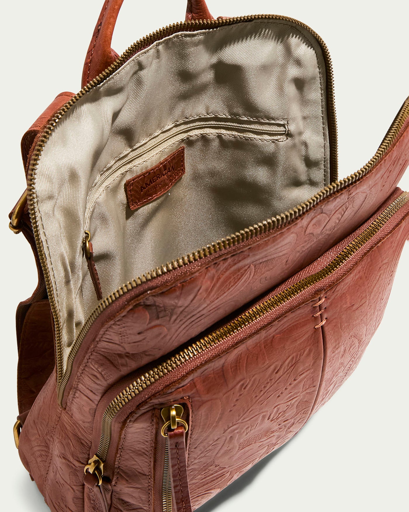 The Cleveland Backpack by American Leather Co. is a brown, genuine leather backpack with intricate patterns and antiqued hardware. Displayed open, it reveals its beige fabric lining, brass zippers, and a front pocket that showcases the interior craftsmanship of this exquisite purse.