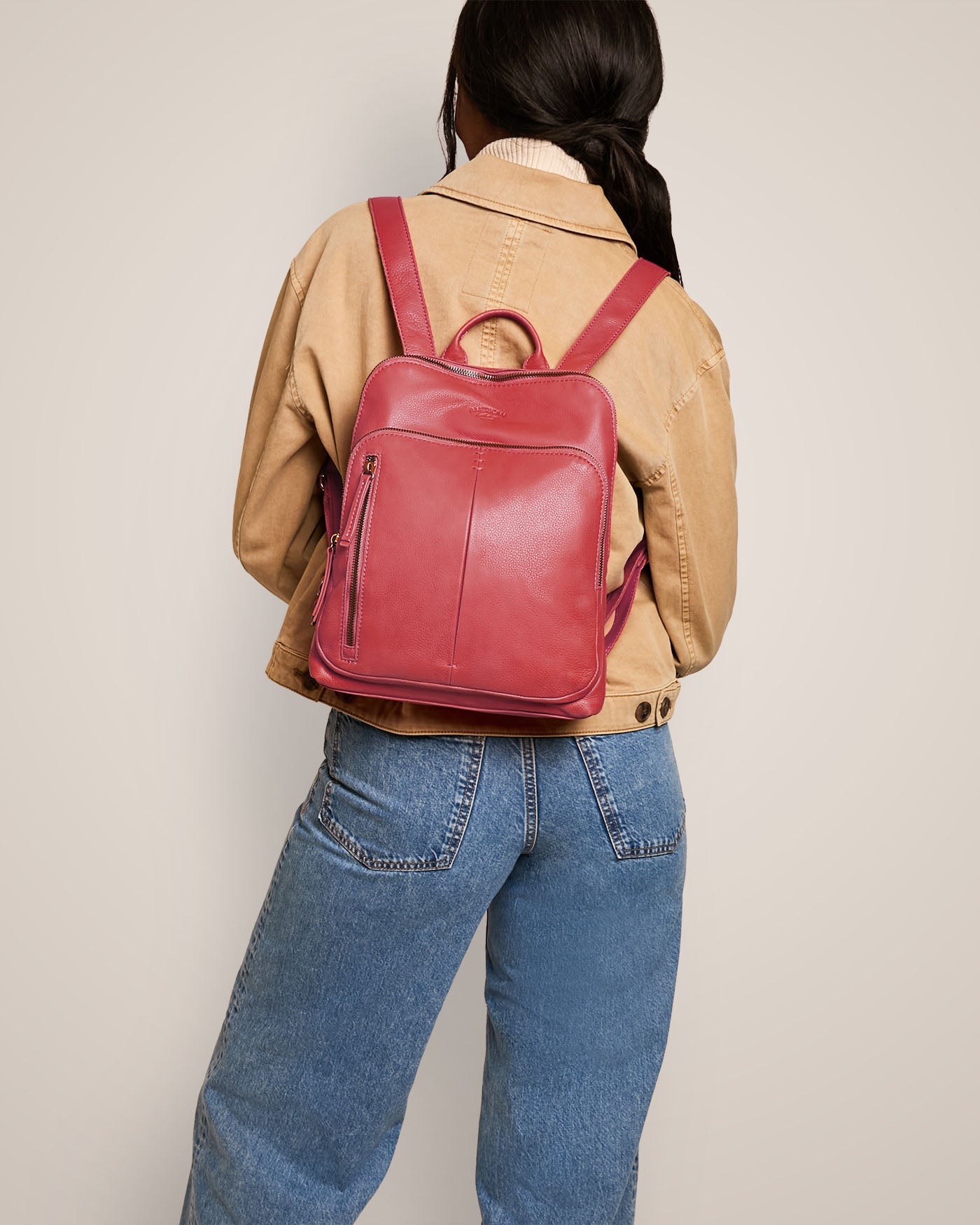 A person with long dark hair is seen from behind, wearing a light brown jacket and blue jeans. They are carrying the Cleveland Backpack by American Leather Co., featuring adjustable back straps, which stands out in red genuine leather against the plain background.