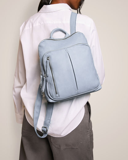 A person in a white shirt and gray pants carries the Cleveland Backpack by American Leather Co., a light blue genuine leather backpack featuring a handle, zippered front pocket, and adjustable back straps.