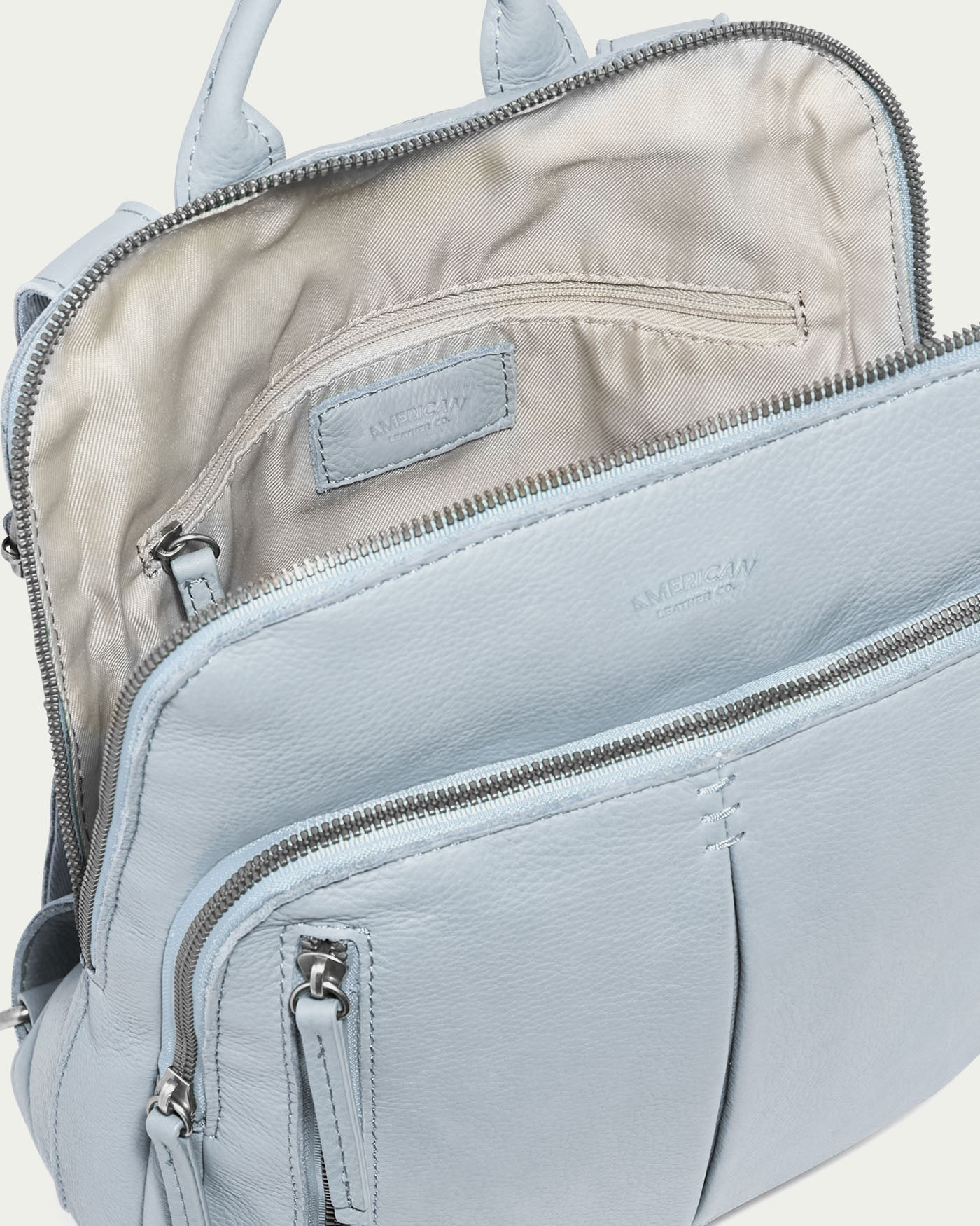 The Cleveland Backpack by American Leather Co. is a light blue genuine leather backpack featuring adjustable back straps, open front and partially open main compartments, a fabric-lined interior with a zip pocket, stitched brand name, metal zippers, and a top handle.